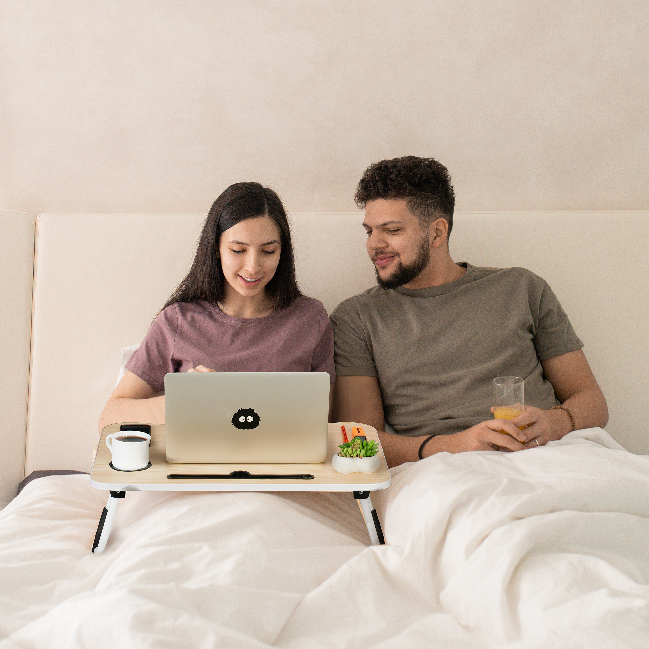 Ein Paar sitzt im Bett; sie arbeitet an einem Laptop auf einem Holztisch, er hält ein Glas Orangensaft.