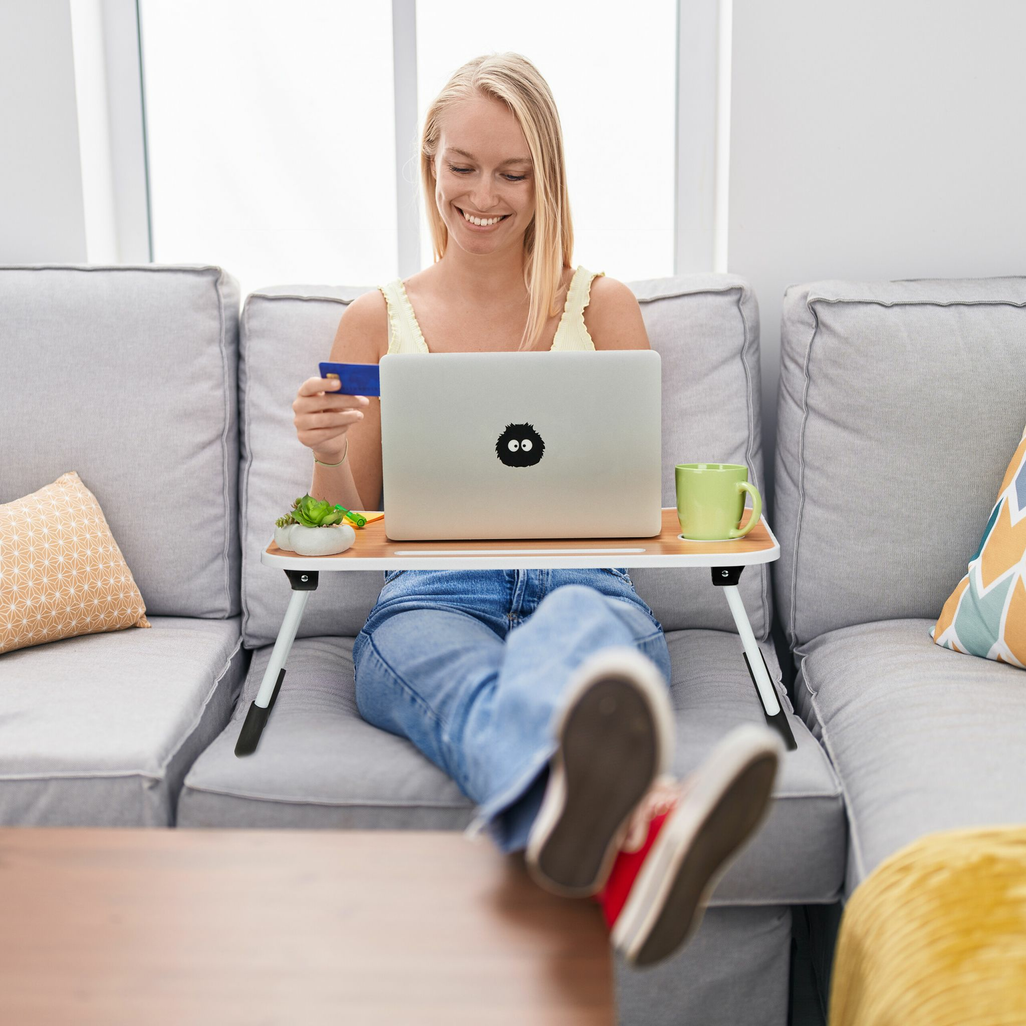 Blonde Frau sitzt auf einem grauen Sofa und nutzt einen Laptop auf einem Schreibtisch, während sie eine blaue Kreditkarte hält.