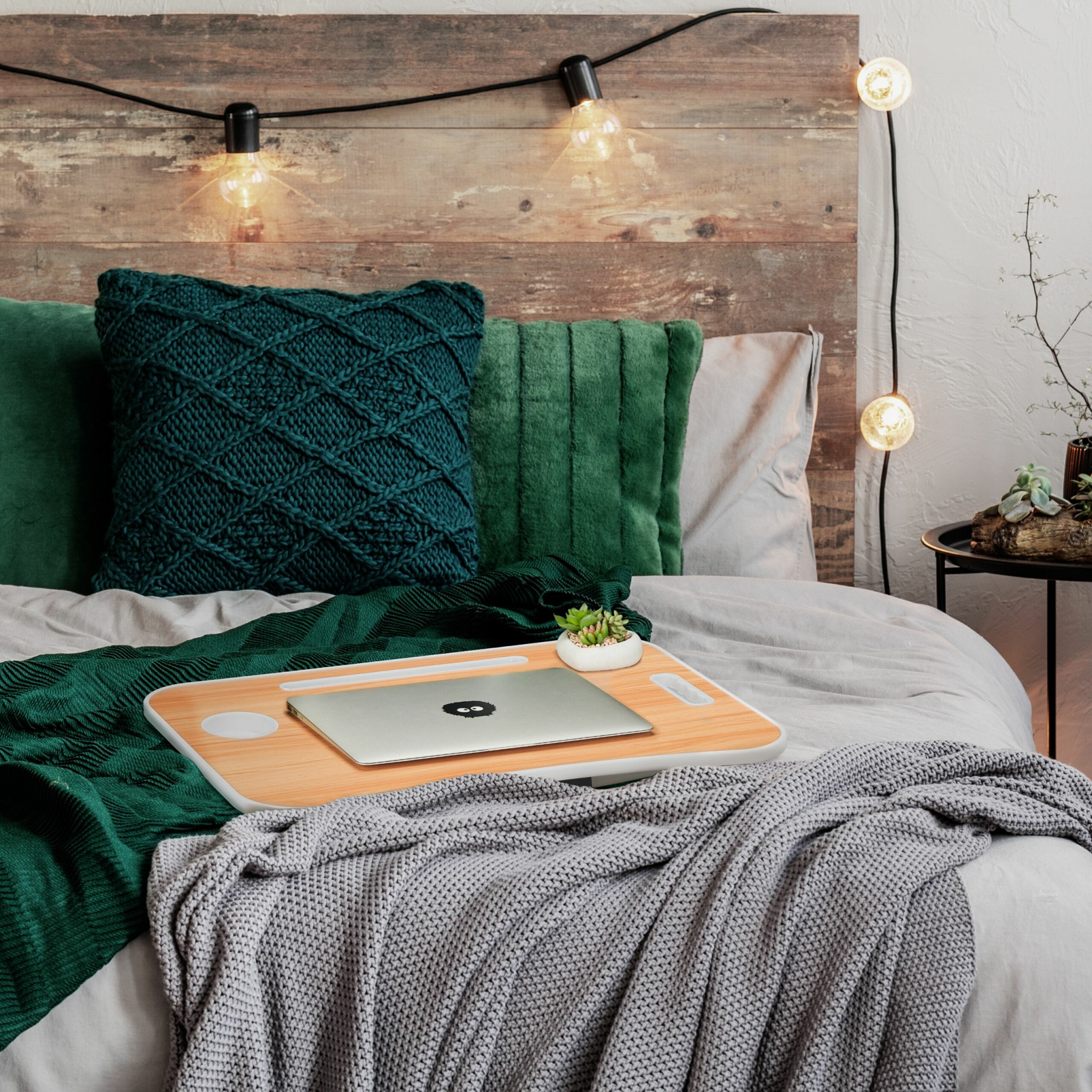 Ein Laptop auf einem Holztablett mit Pflanze, umgeben von grünen Kissen und Decken auf einem Bett.