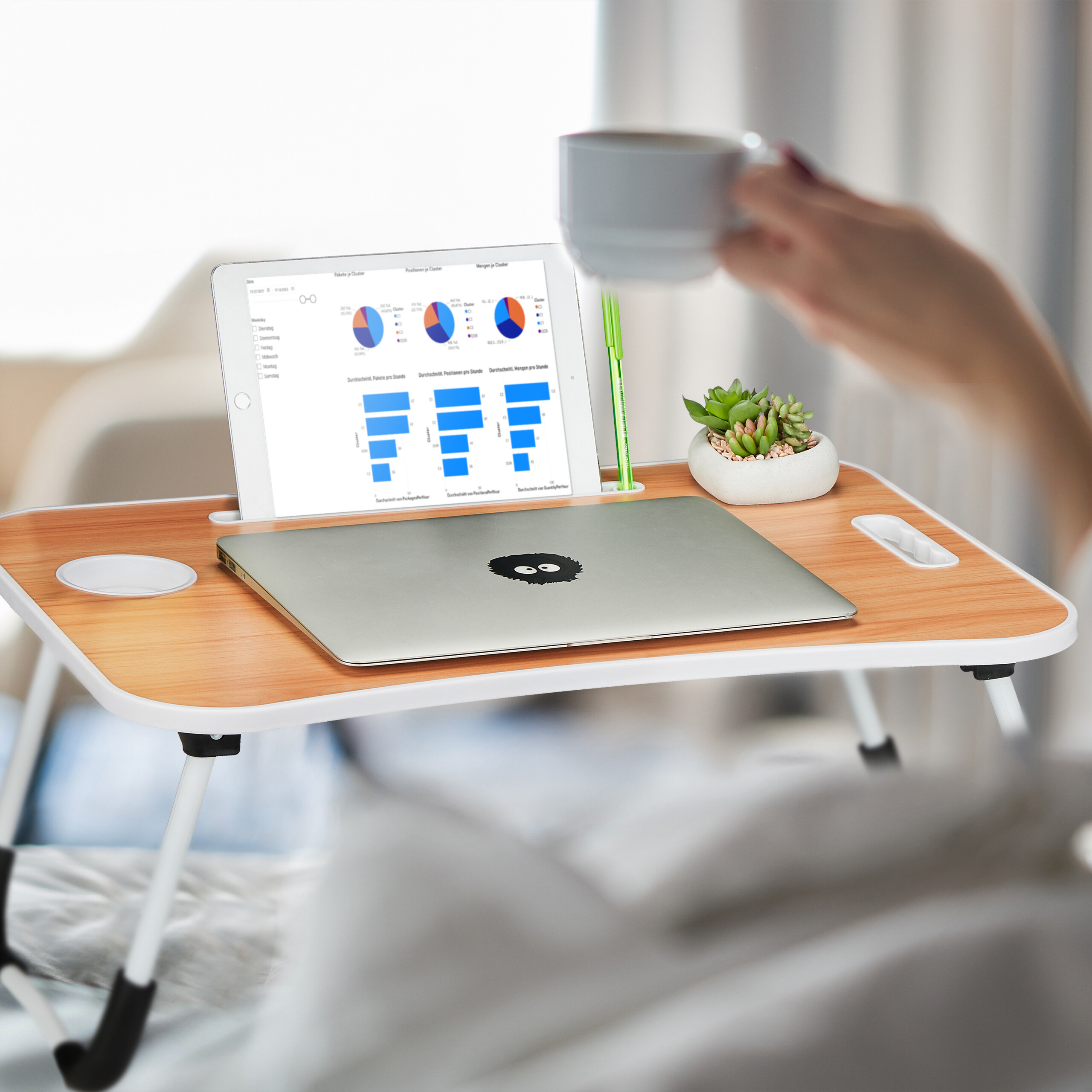 Ein Holzbett-Tablett mit einem Laptop, einem Tablet mit Diagrammen, einem kleinen Sukkulenten-Topf und einer Hand mit Tasse.