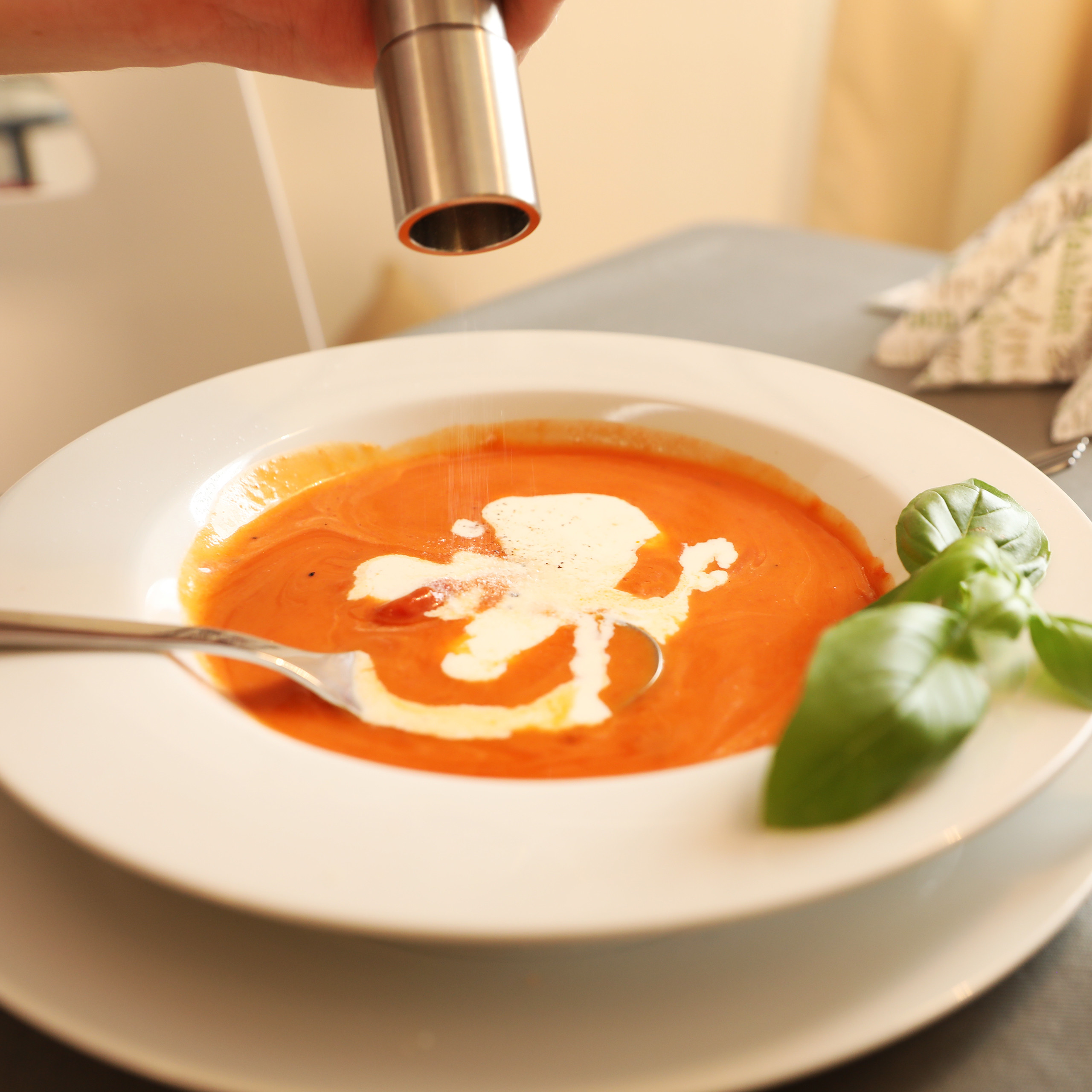 Eine Hand würzt Tomatensuppe mit Sahne und Basilikum in einem weißen Teller.