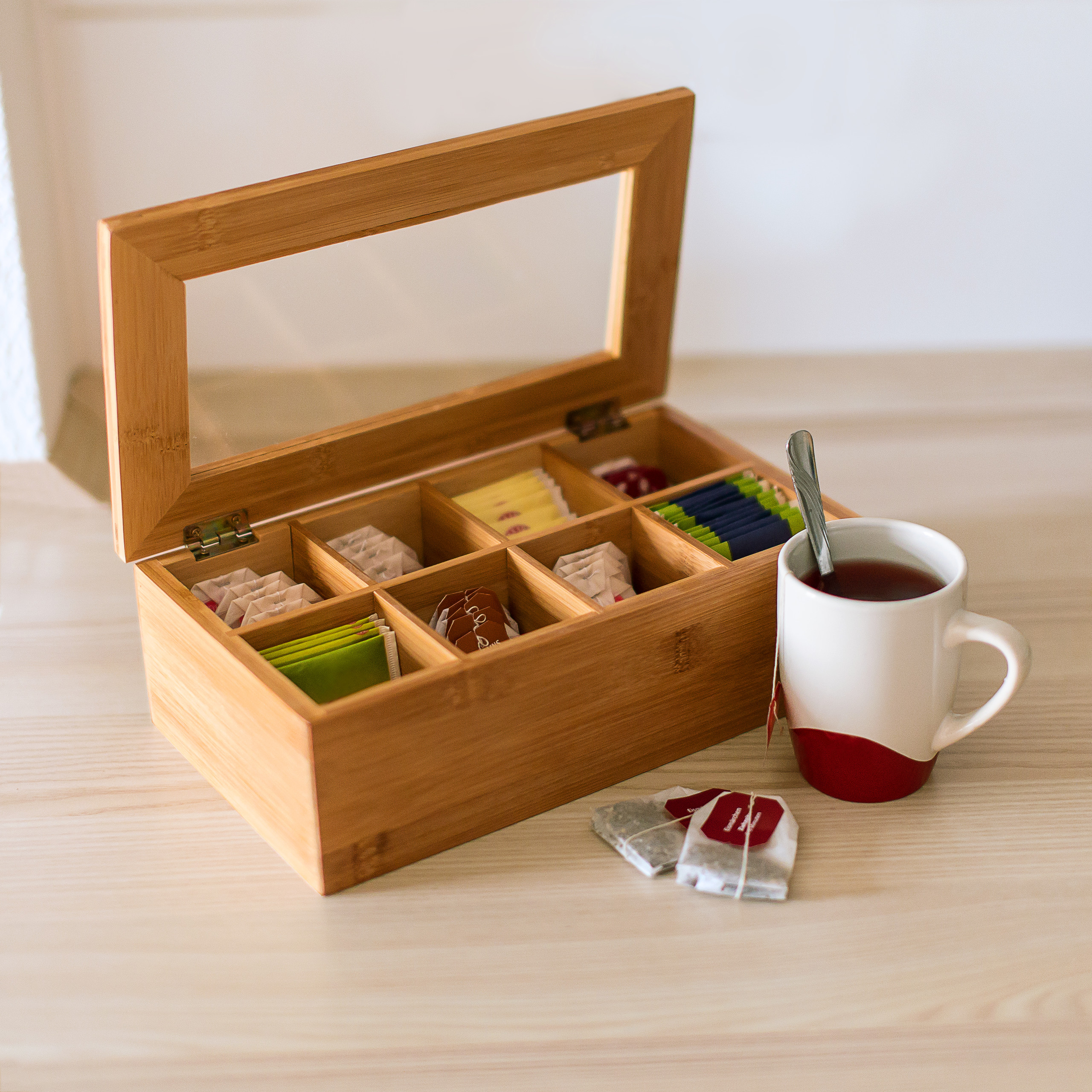 Eine offene Teebox aus Holz mit verschiedenen Teebeuteln und eine Tasse Tee mit Löffel stehen auf dem Tisch.