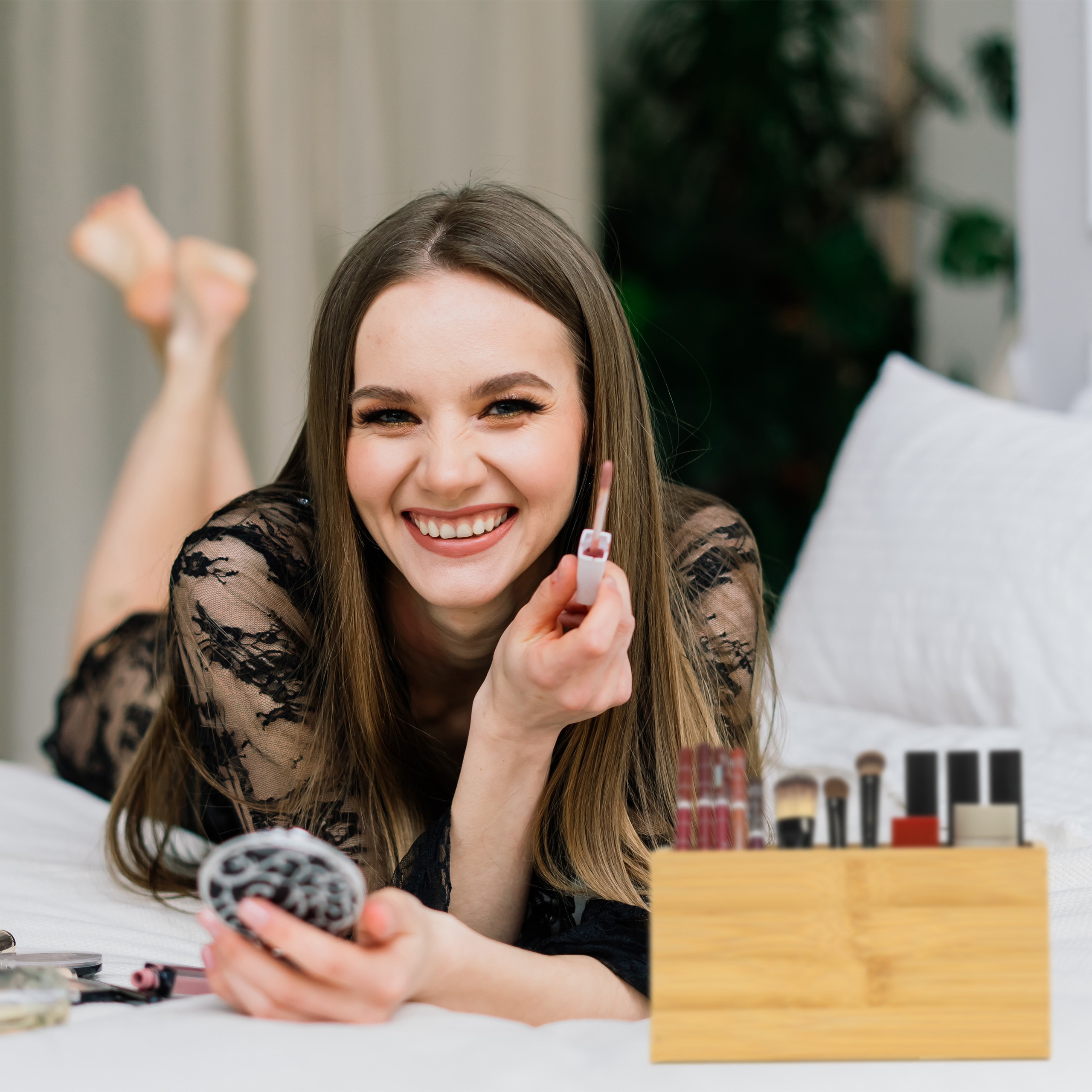 Lächelnte Frau in schwarzer Spitze auf dem Bett, hält Lipgloss neben Make-up-Organizer.