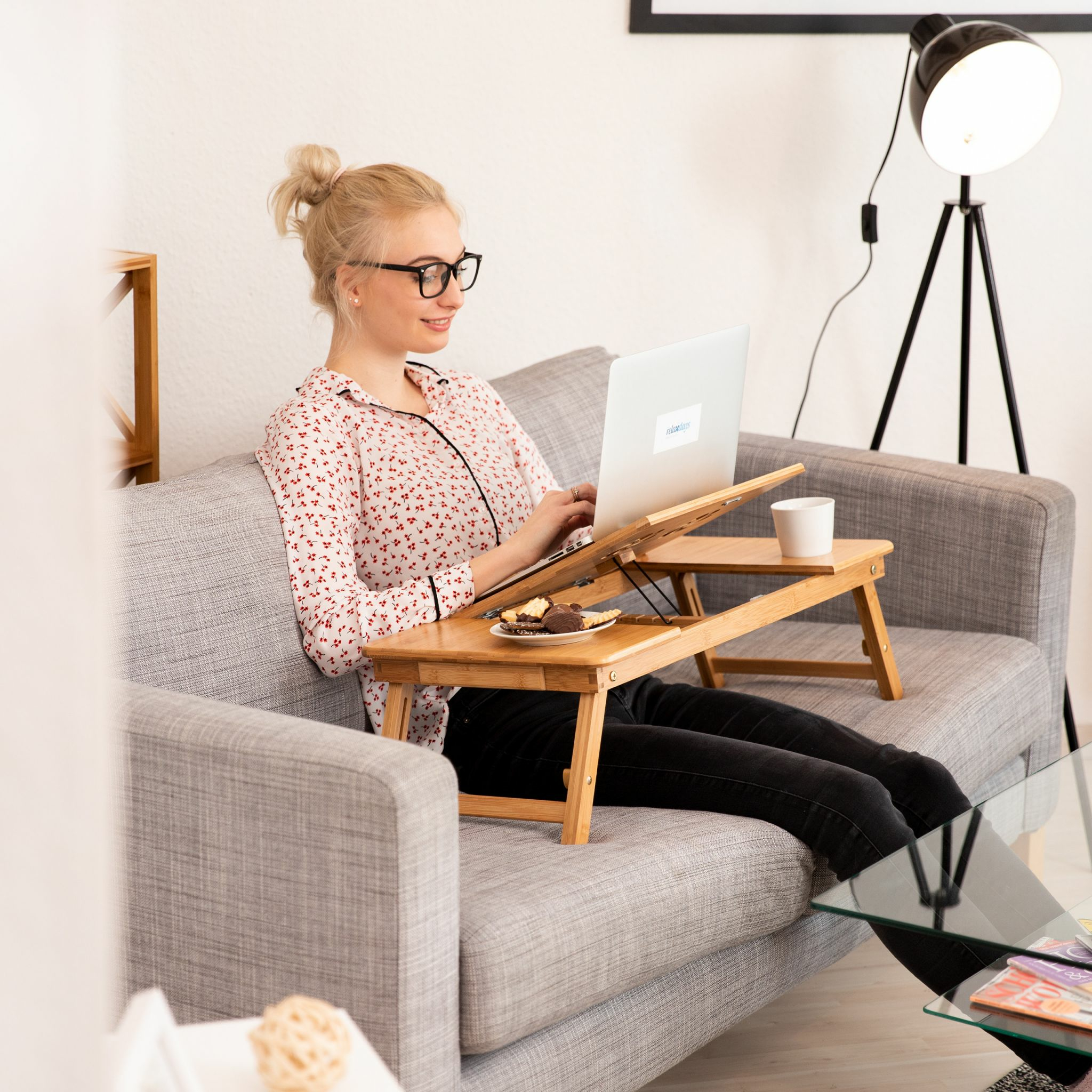 Eine blonde Frau sitzt auf einem grauen Sofa und arbeitet an einem Laptop auf einem Holztisch.