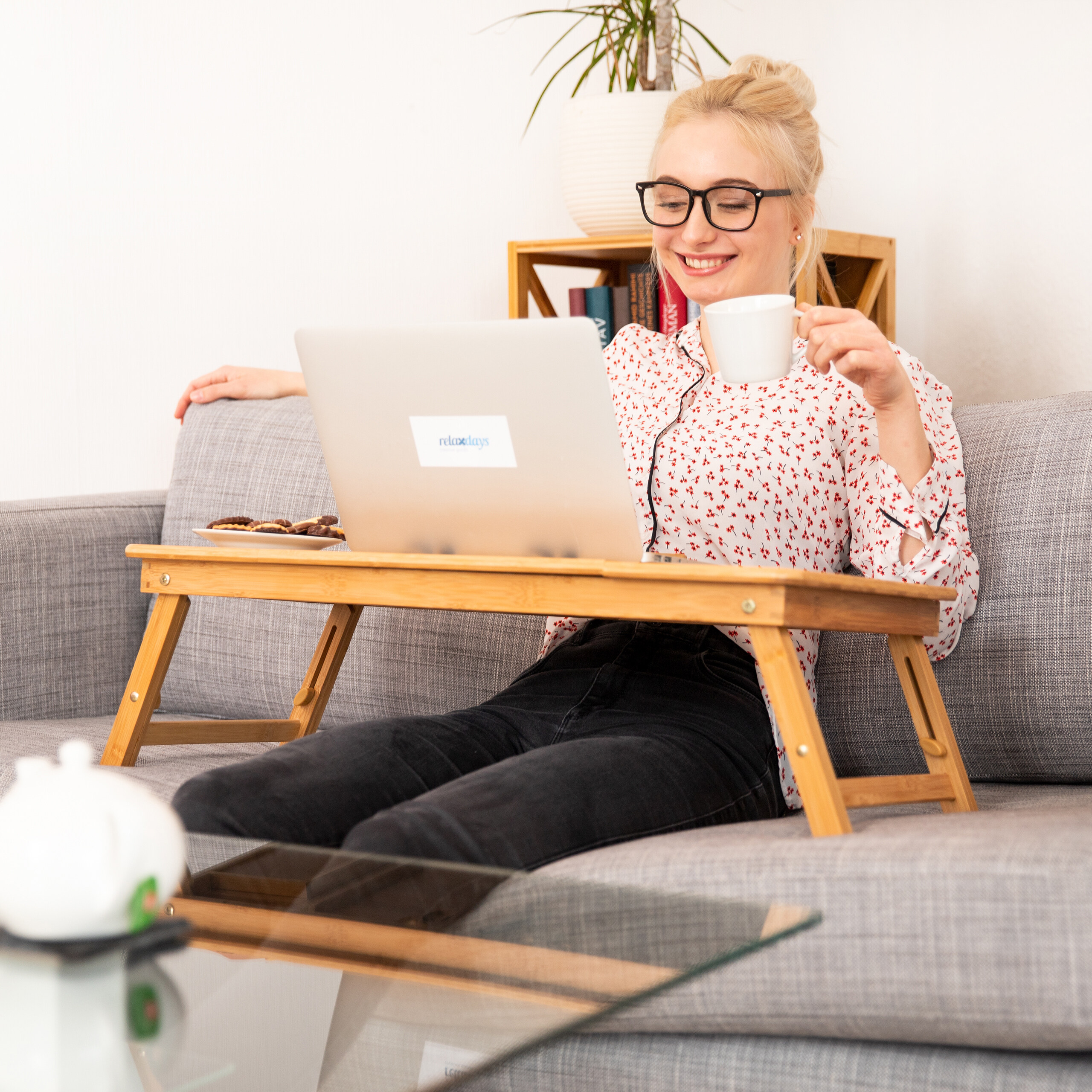 Eine blonde Frau mit Brille sitzt auf einem grauen Sofa, hält eine weiße Tasse und arbeitet am Laptop auf einem Holztisch.