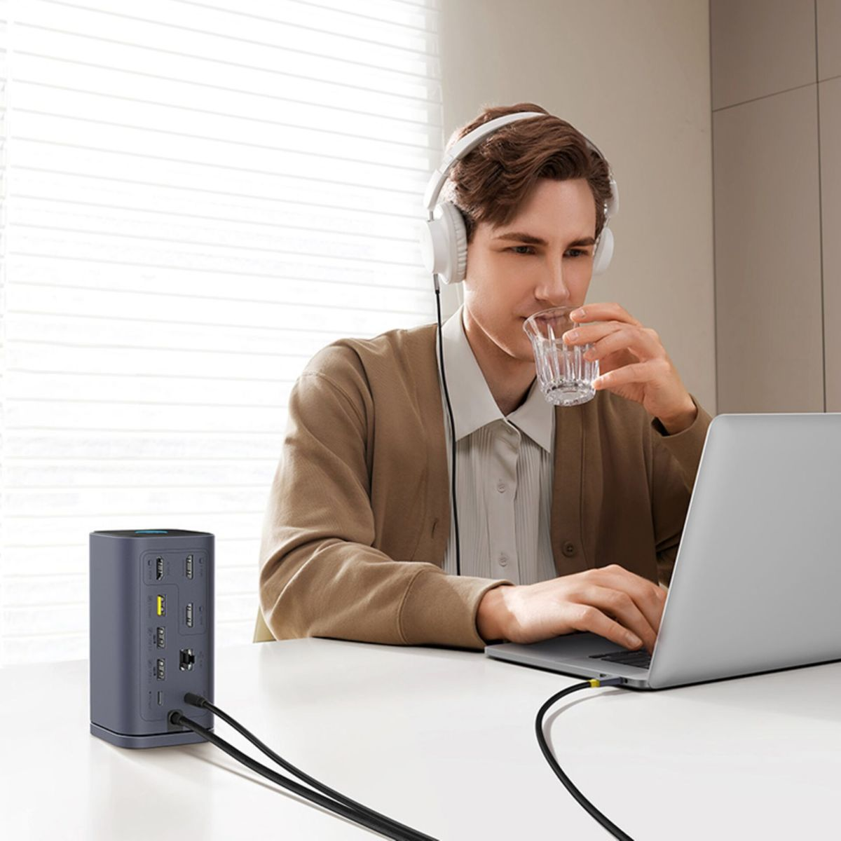 Ein Mann mit Kopfhörern trinkt Wasser, während er am Laptop arbeitet, neben einer grauen Dockingstation.