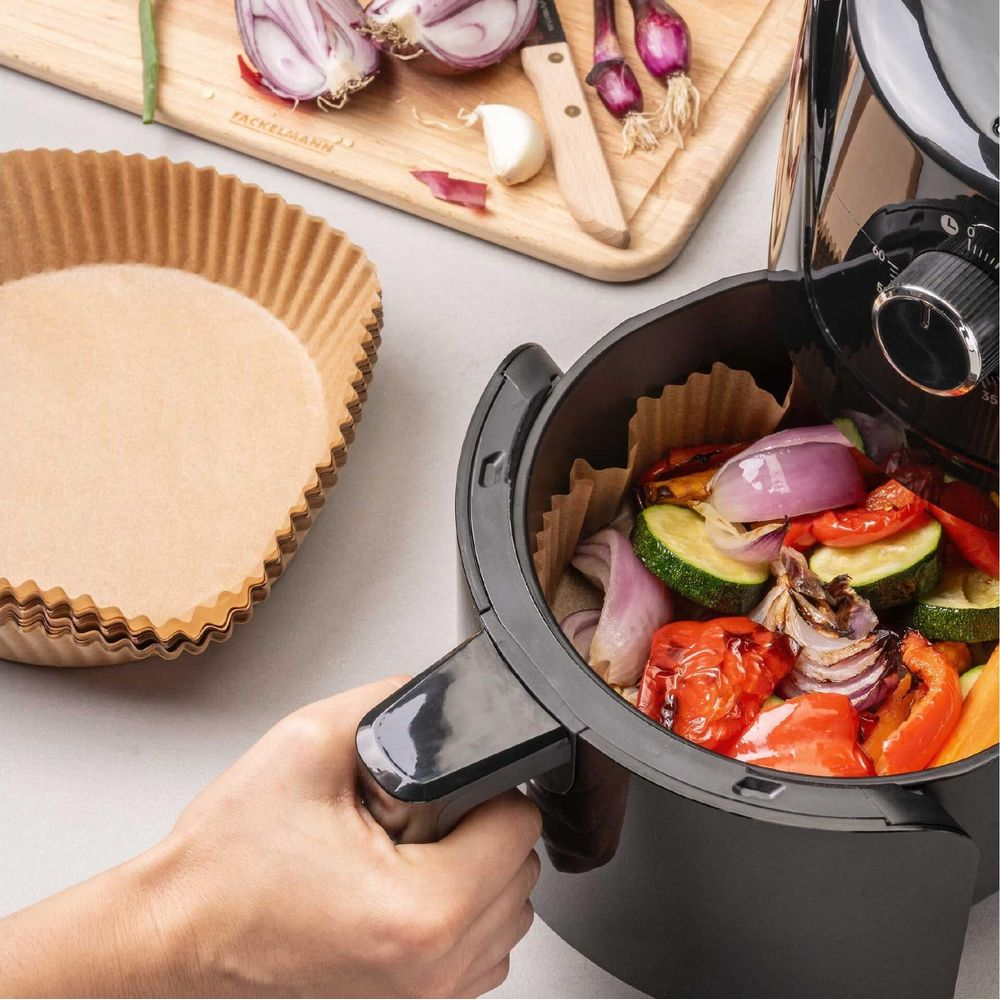 Mano regge friggitrice con verdure, carta da forno e tagliere vicini.