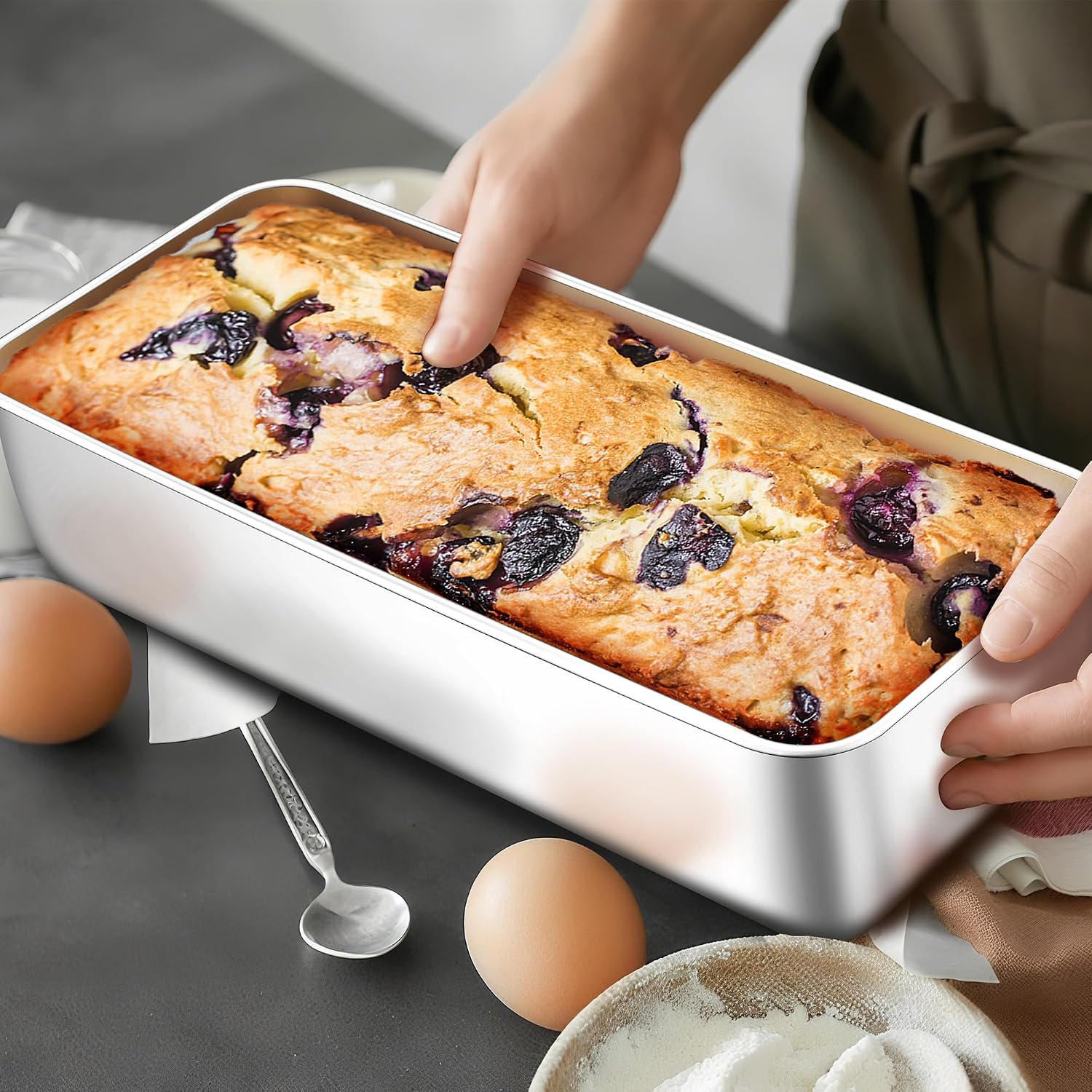 Eine Person hält eine frische Blaubeerkuchen-Kastenform auf einer Arbeitsplatte mit Eiern und Mehl.