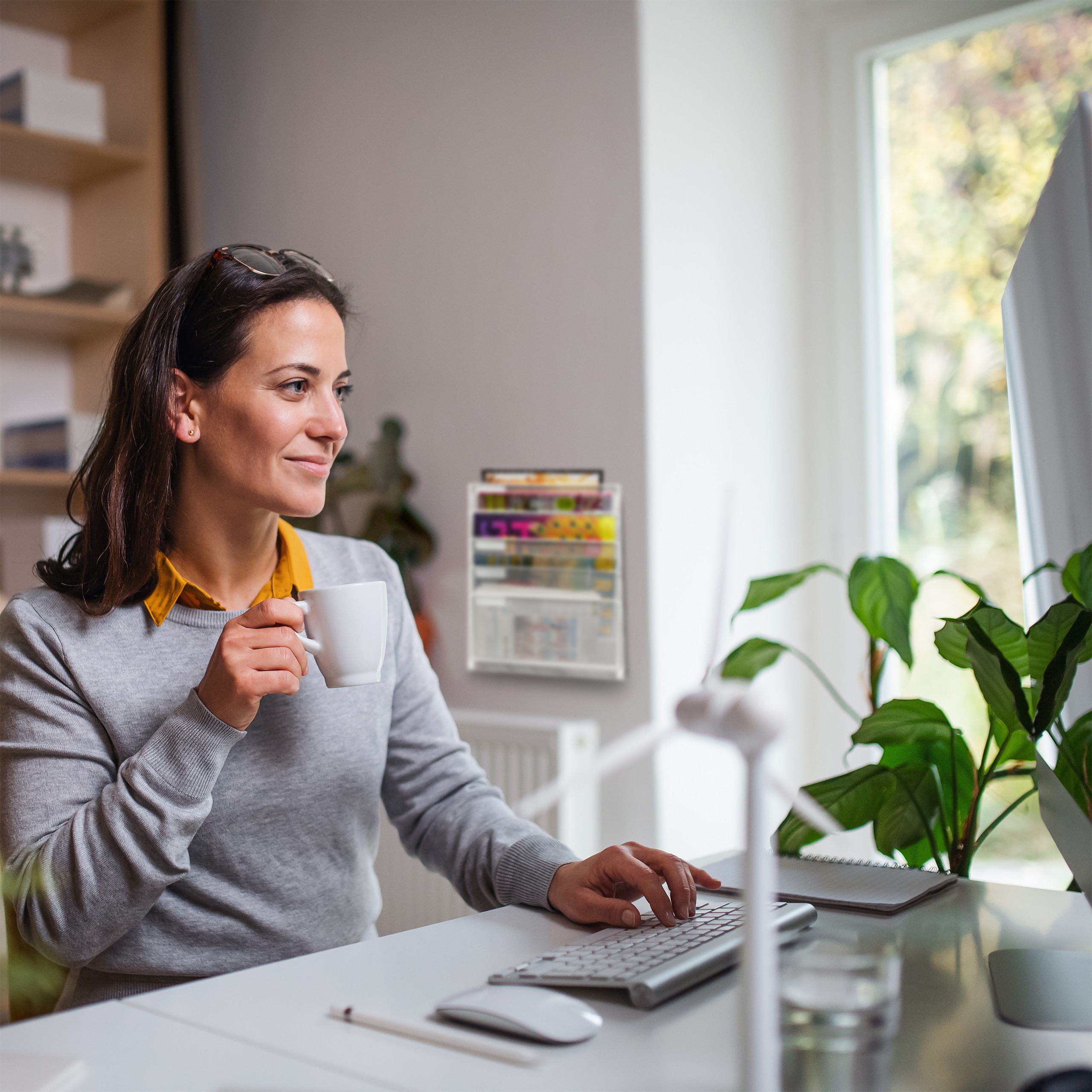 Eine Frau im Grauen Pullover trinkt Kaffee und arbeitet am Computer in einem hellen Büro.