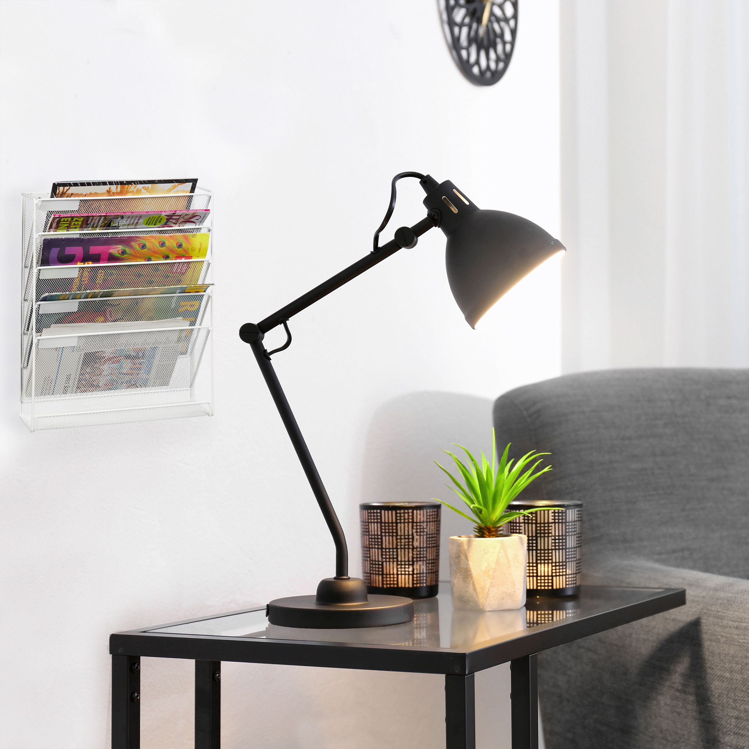 Schwarze Schreibtischlampe, kleine Pflanze und Kerzenhalter auf Glastisch vor weißer Wand mit Zeitschriftenhalter.