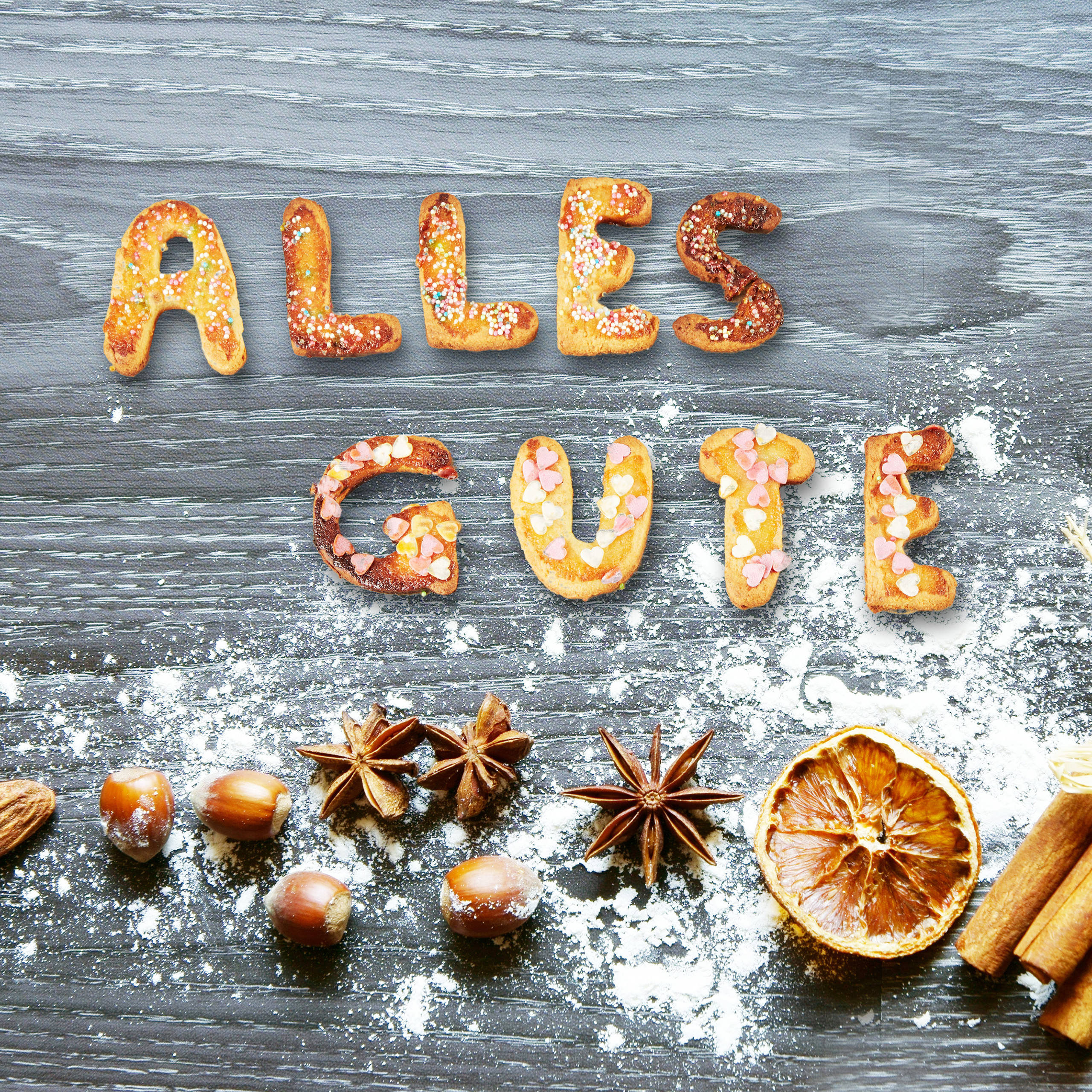 Kekse mit der Aufschrift Alles Gute, umgeben von Nüssen, Sternanis, Orangenscheiben und Puderzucker.