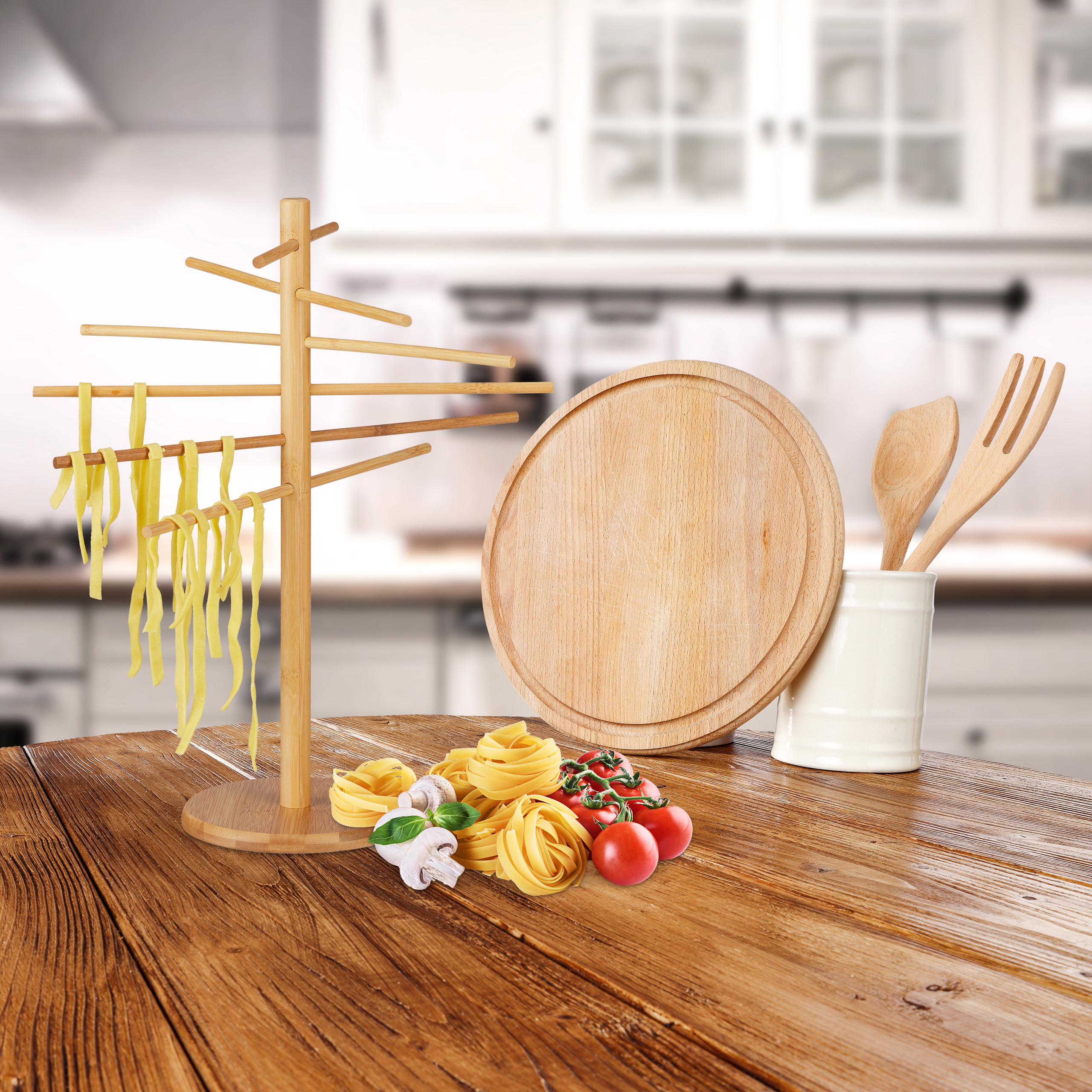Holztisch mit Pastaständer, Schneidebrett, Tomaten und Pilzen vor einer unscharfen Küche.