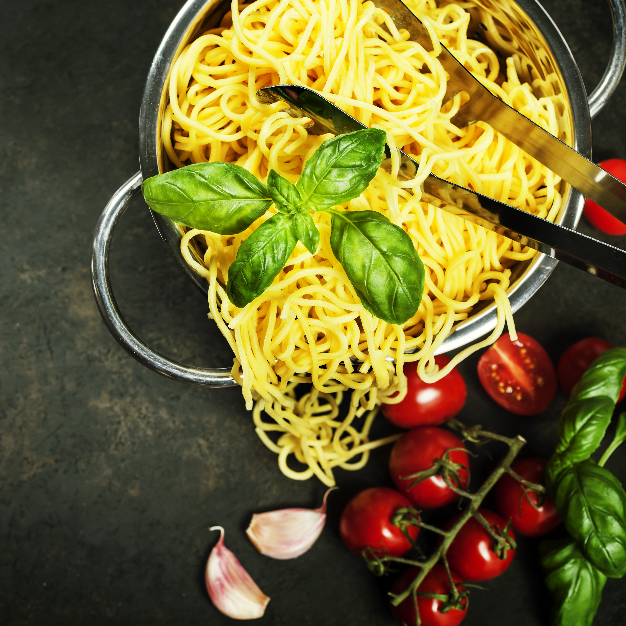 Gekochte Spaghetti in einem Sieb, garniert mit frischem Basilikum, Tomaten und Knoblauch auf dunklem Hintergrund.