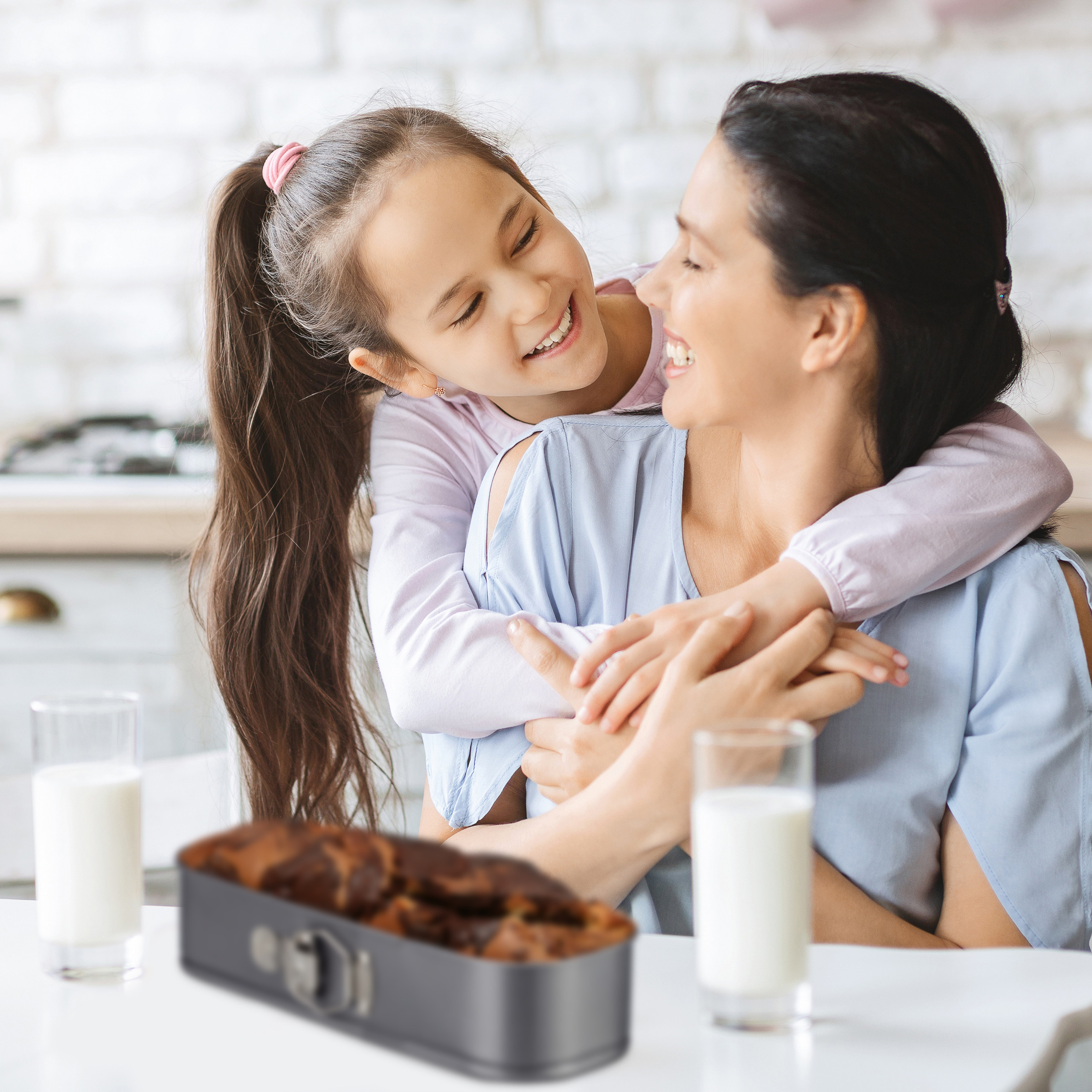 Ein junges Mädchen lächelt und umarmt ihre Mutter in einer hellen Küche mit Gläsern voll Milch und Kuchen.