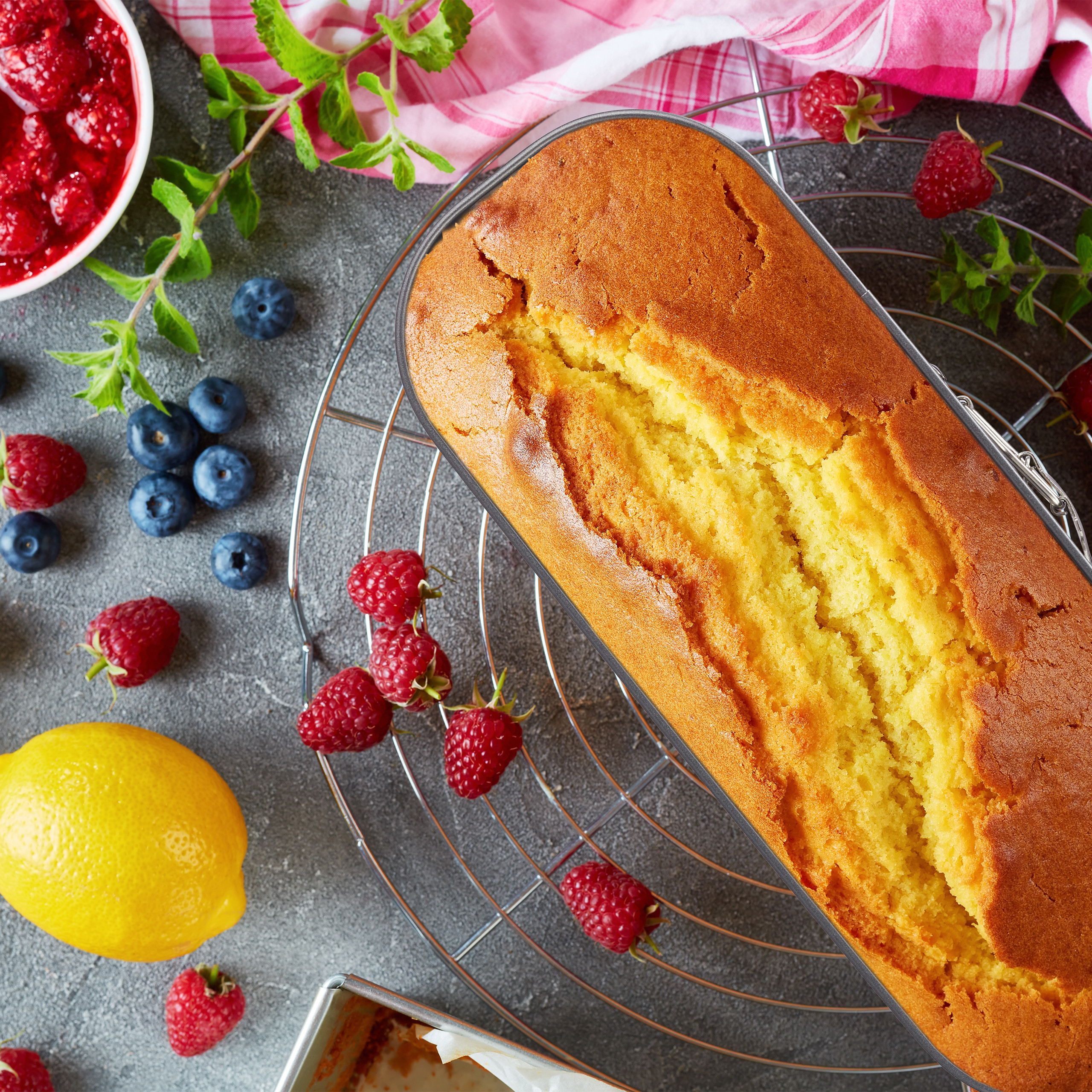 Goldbrauner Kuchen auf Kuchengitter mit Zitronen, Himbeeren und Blaubeeren auf dunklem Stein.
