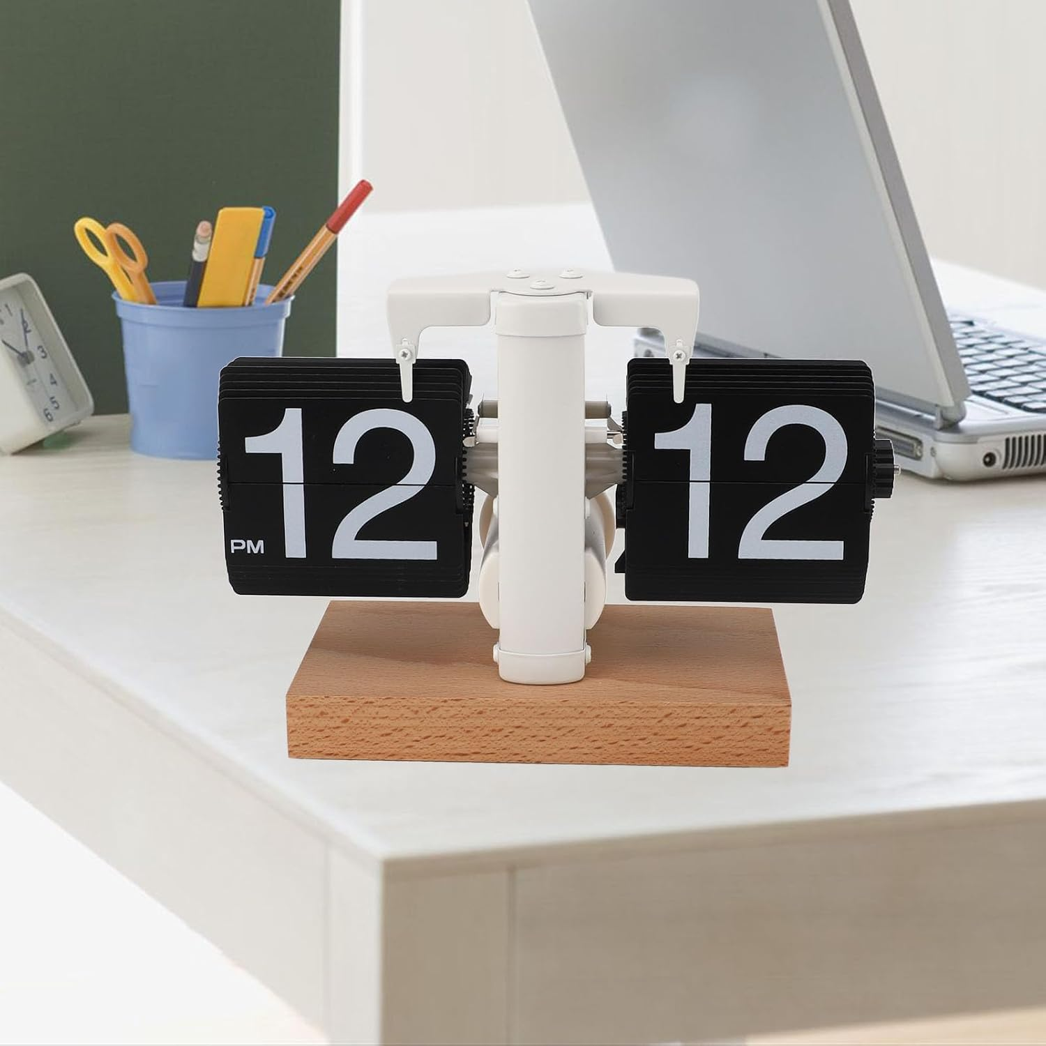 Ein weißer Flip-Uhr mit Holzsockel auf einem Tisch, im Hintergrund ein Laptop und Büromaterial.