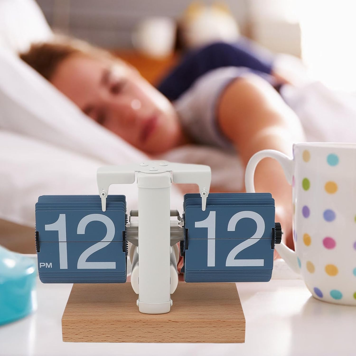 Ein blaues Fallblatt-Wecker zeigt 12:12 Uhr an, im Hintergrund schläft eine Frau in einem Bett.