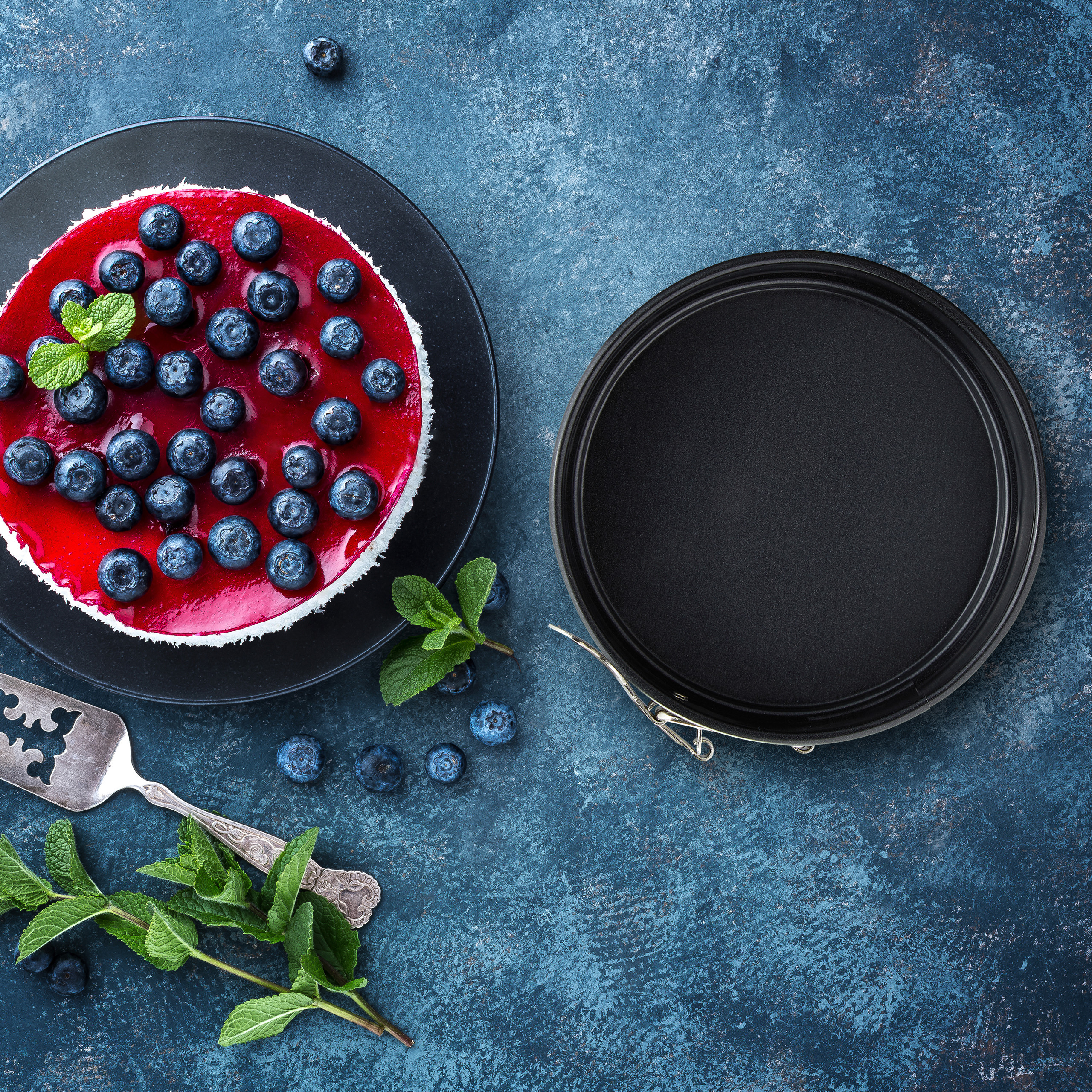 Ein Blaubeer-Käsekuchen auf einem Teller, eine Springform und Minzblätter auf blauem Untergrund.