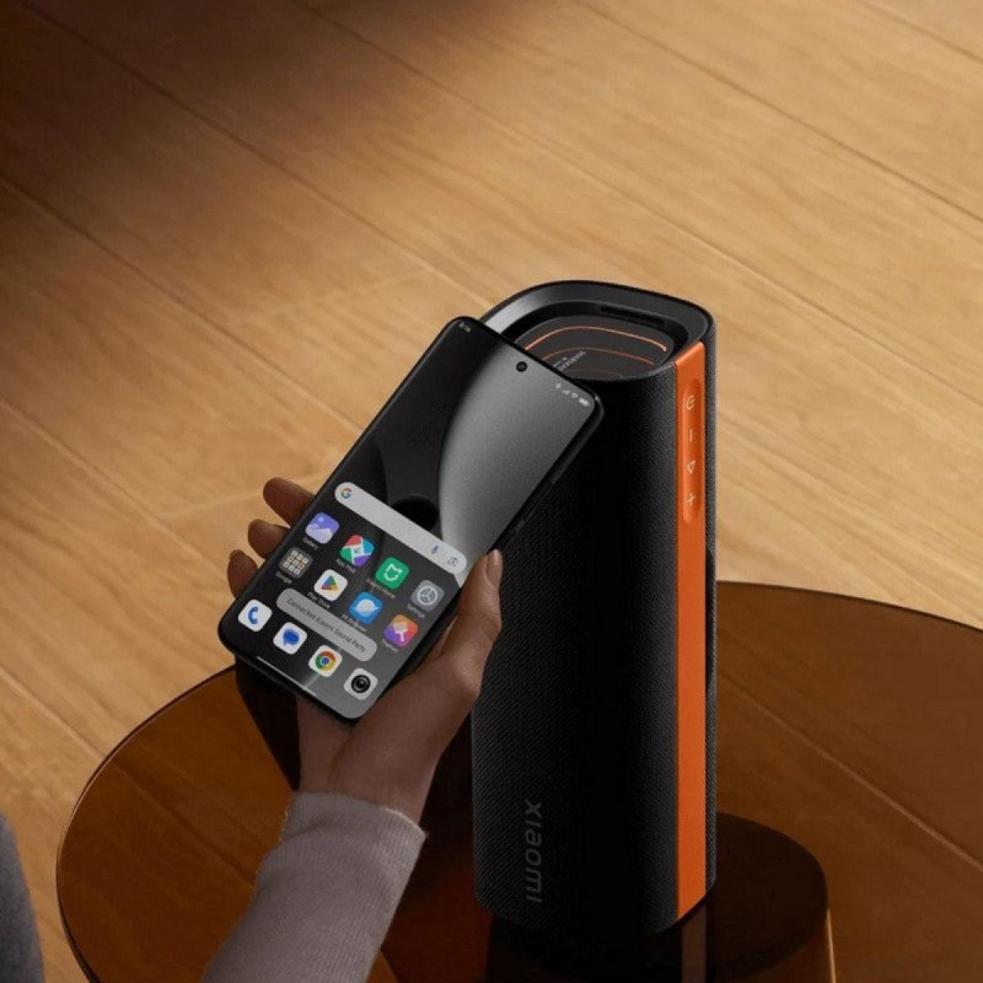 Una mano tiene uno smartphone sopra un altoparlante cilindrico nero e arancione Xiaomi su un tavolo di legno.
