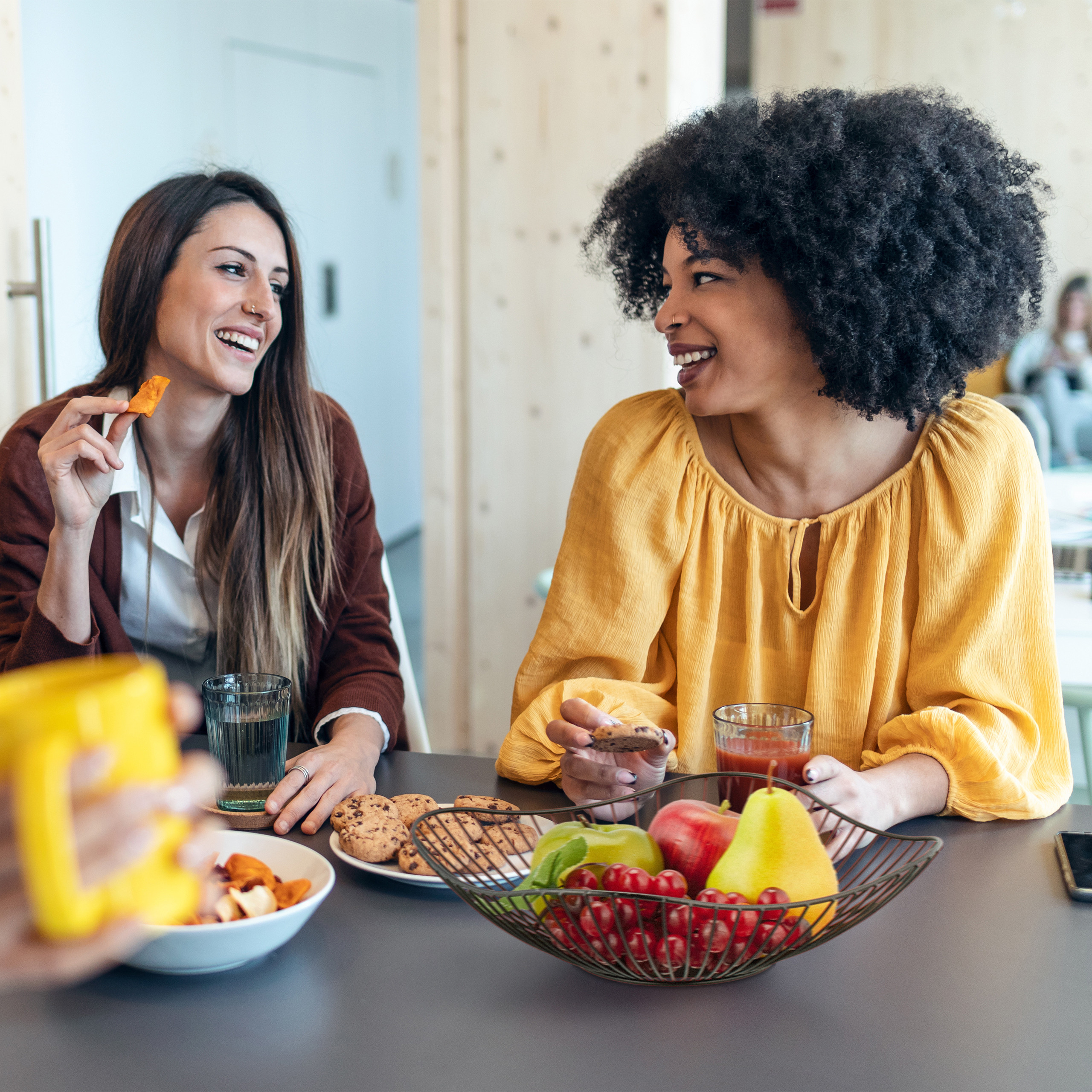 Due donne sorridenti siedono a un tavolo, mangiando biscotti e snack mentre chiacchierano.