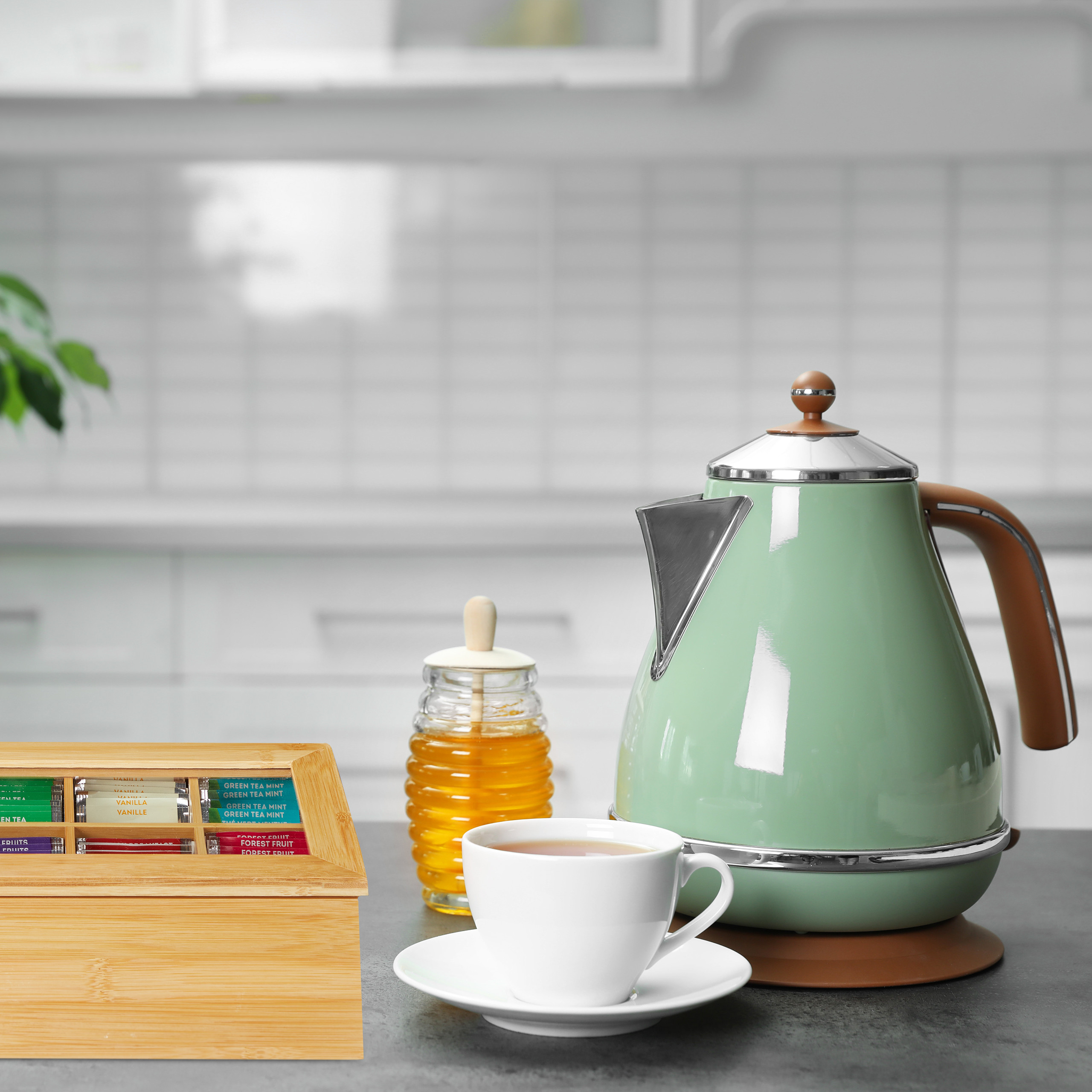 Ein mintgrüner Wasserkocher, eine Tasse Tee, ein Honigglas und eine Teebox auf einer Küchenzeile.
