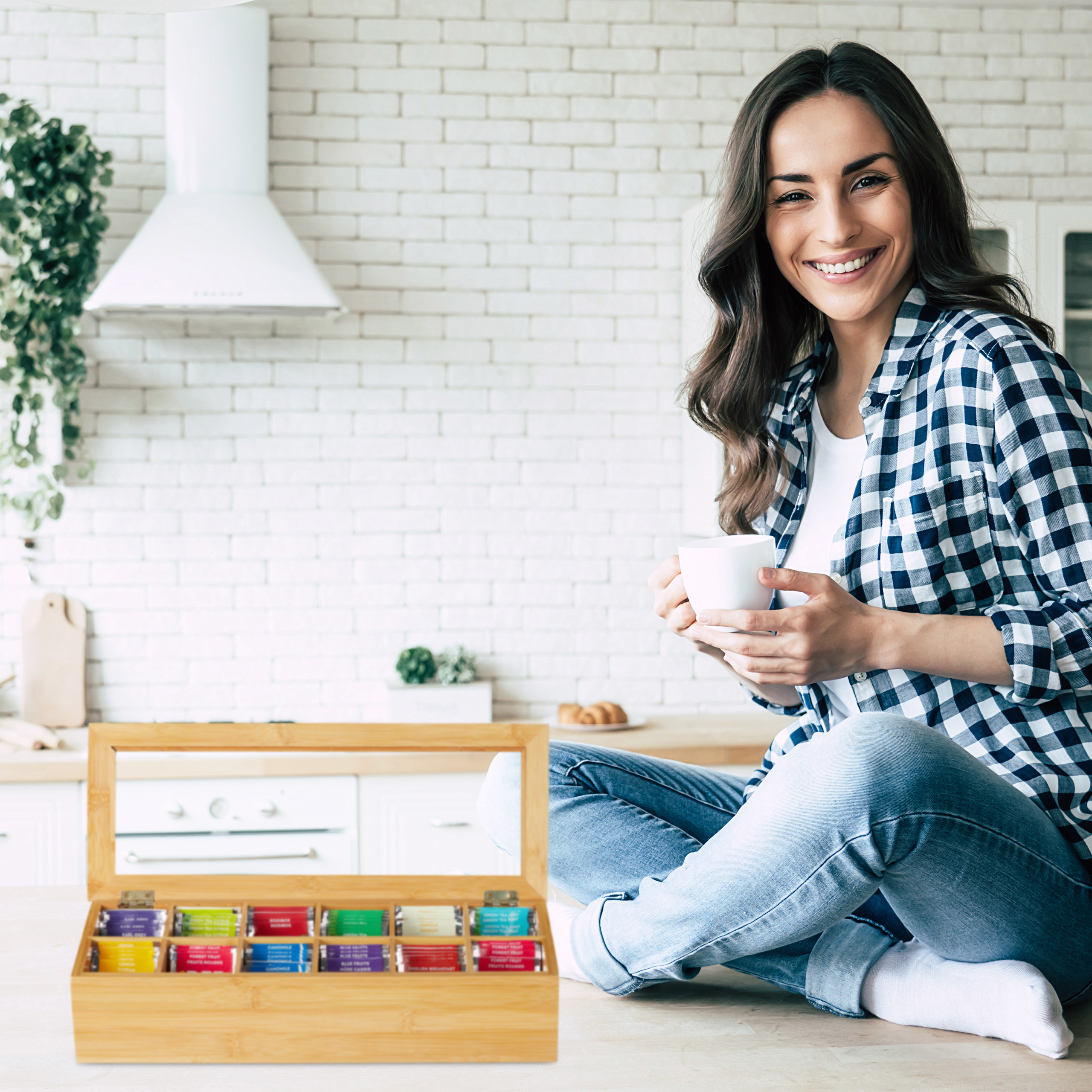 Eine lächelnde Frau sitzt mit einer Tasse in einer Küche neben einer Teebox.