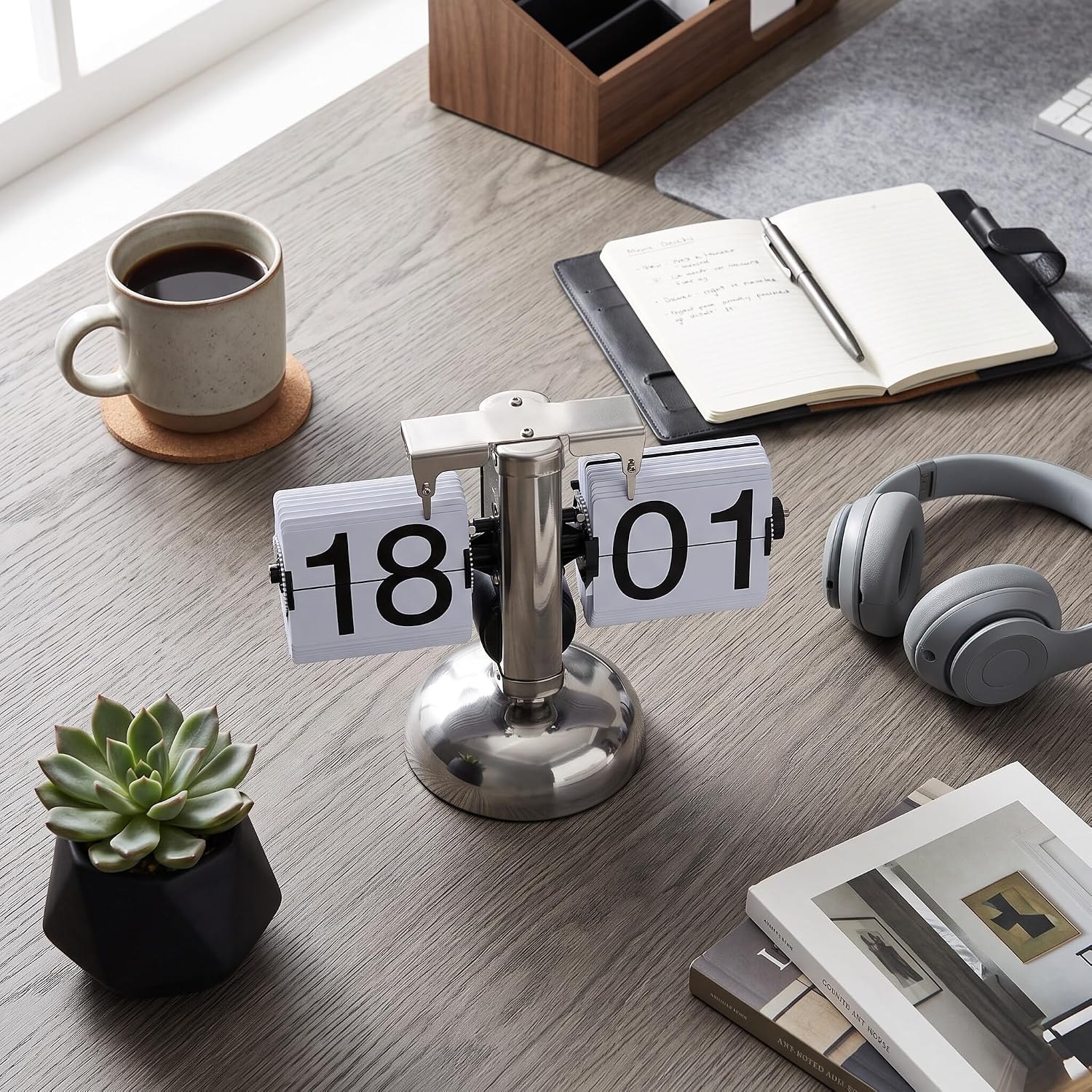 Ein holzfarbener Schreibtisch mit Flip-Uhr, Notizbuch, Kaffeetasse, Kopfhörern, Pflanze und Büchern.