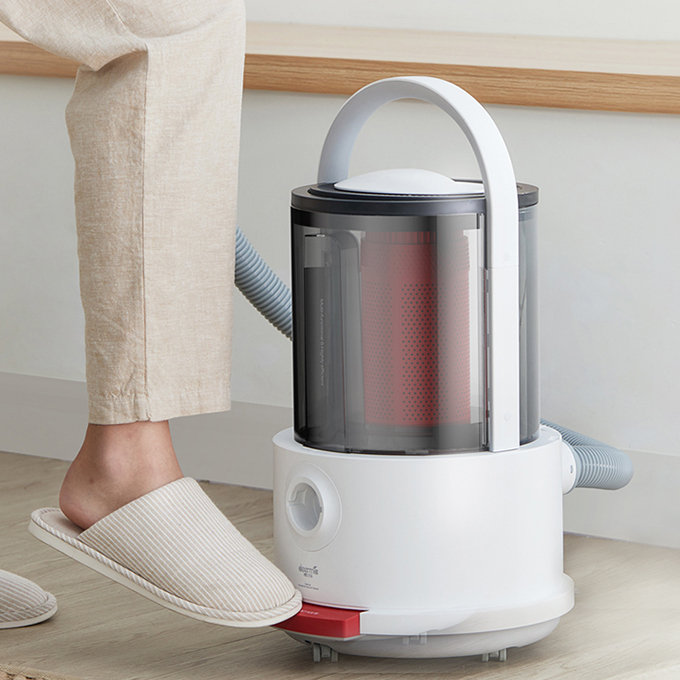 Un piede in pantofola a righe preme il pedale rosso di un aspirapolvere bianco e grigio sul pavimento.