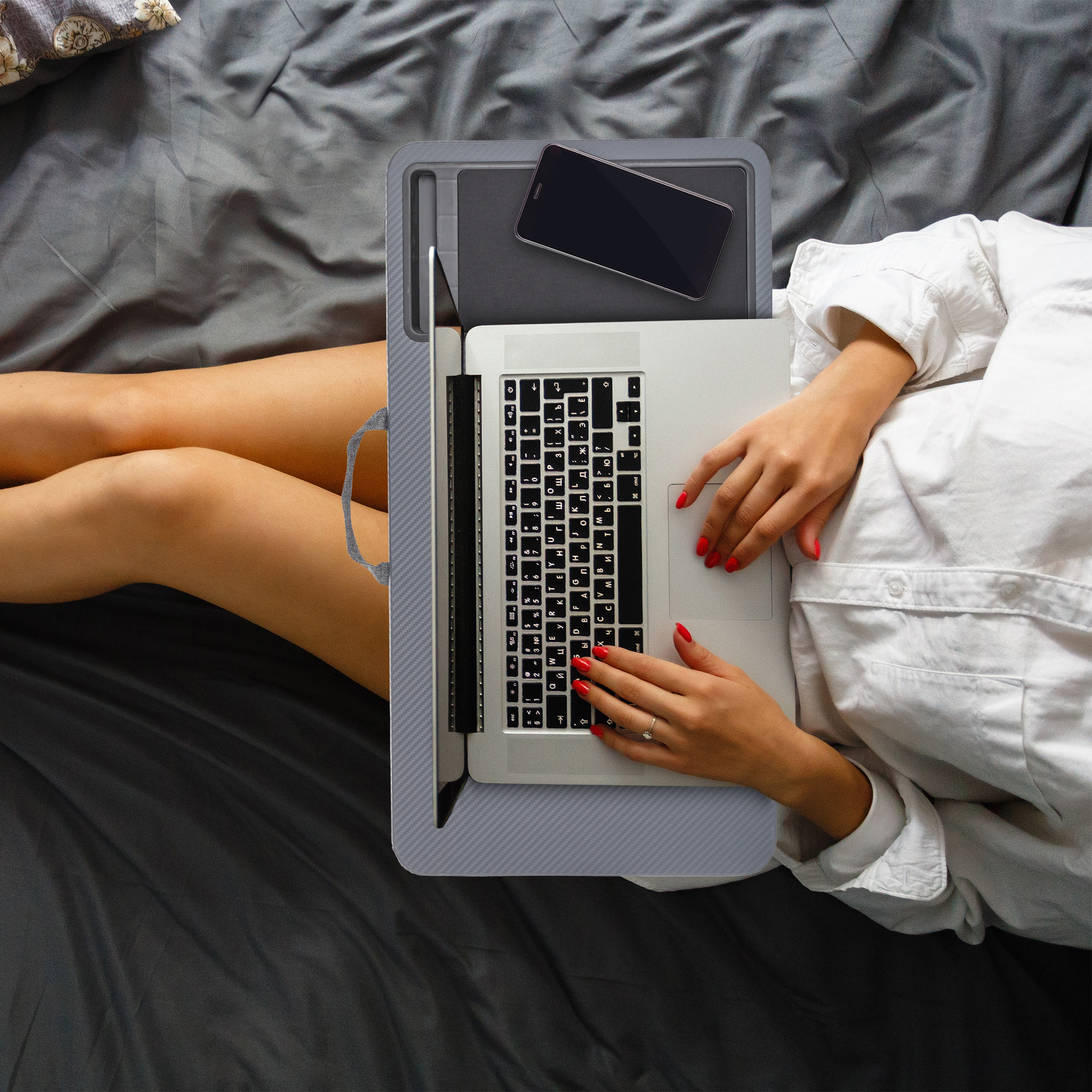 Eine Frau in weißem Hemd nutzt im Bett einen Laptop auf einem Schreibtisch mit Smartphone.