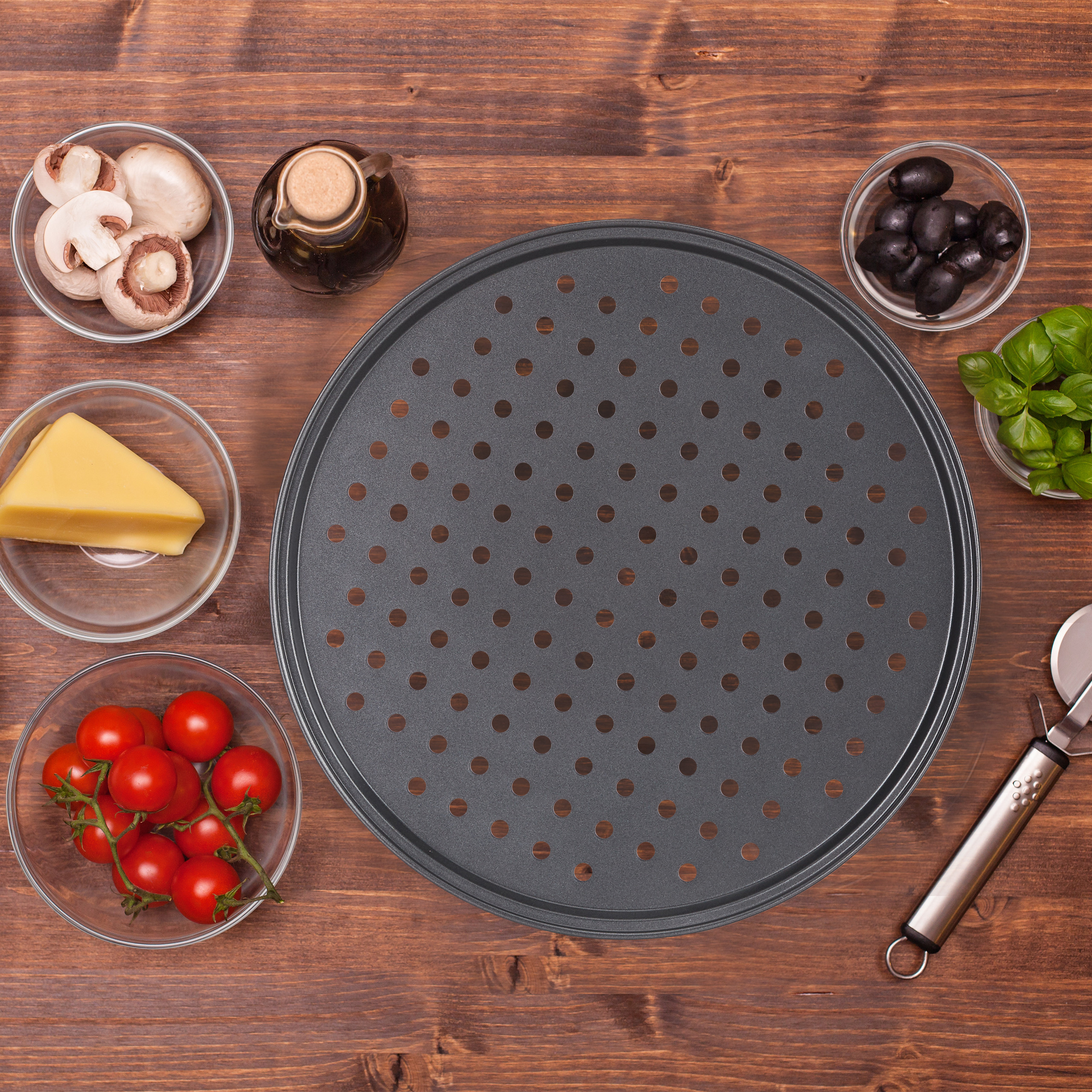 Ein Pizzablech mit Löchern ist von Zutaten wie Tomaten, Pilzen, Käse, Oliven und Basilikum auf Holz umgeben.