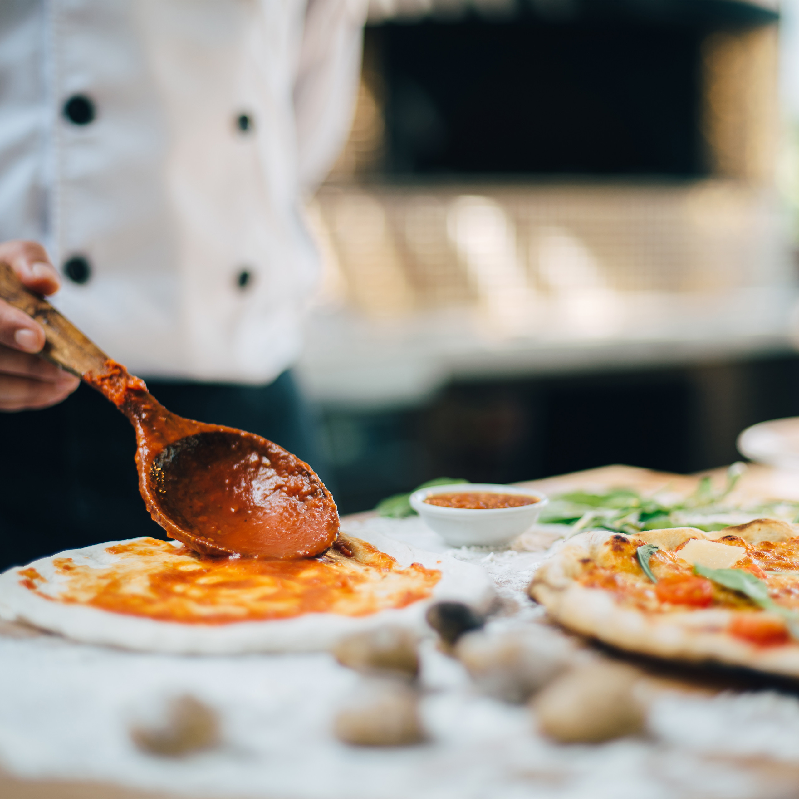 Ein Koch verteilt Tomatensauce mit einem Holzlöffel auf einem rohen Pizzateig.