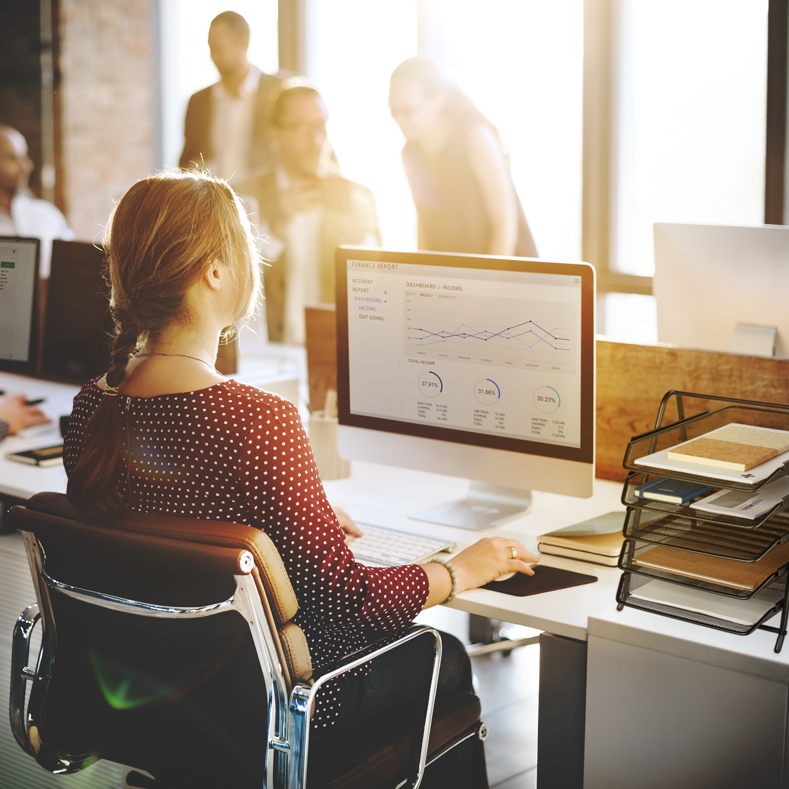 Eine Frau sitzt am Schreibtisch vor einem Computer mit Finanzdiagrammen, im Hintergrund sind Kollegen zu sehen.