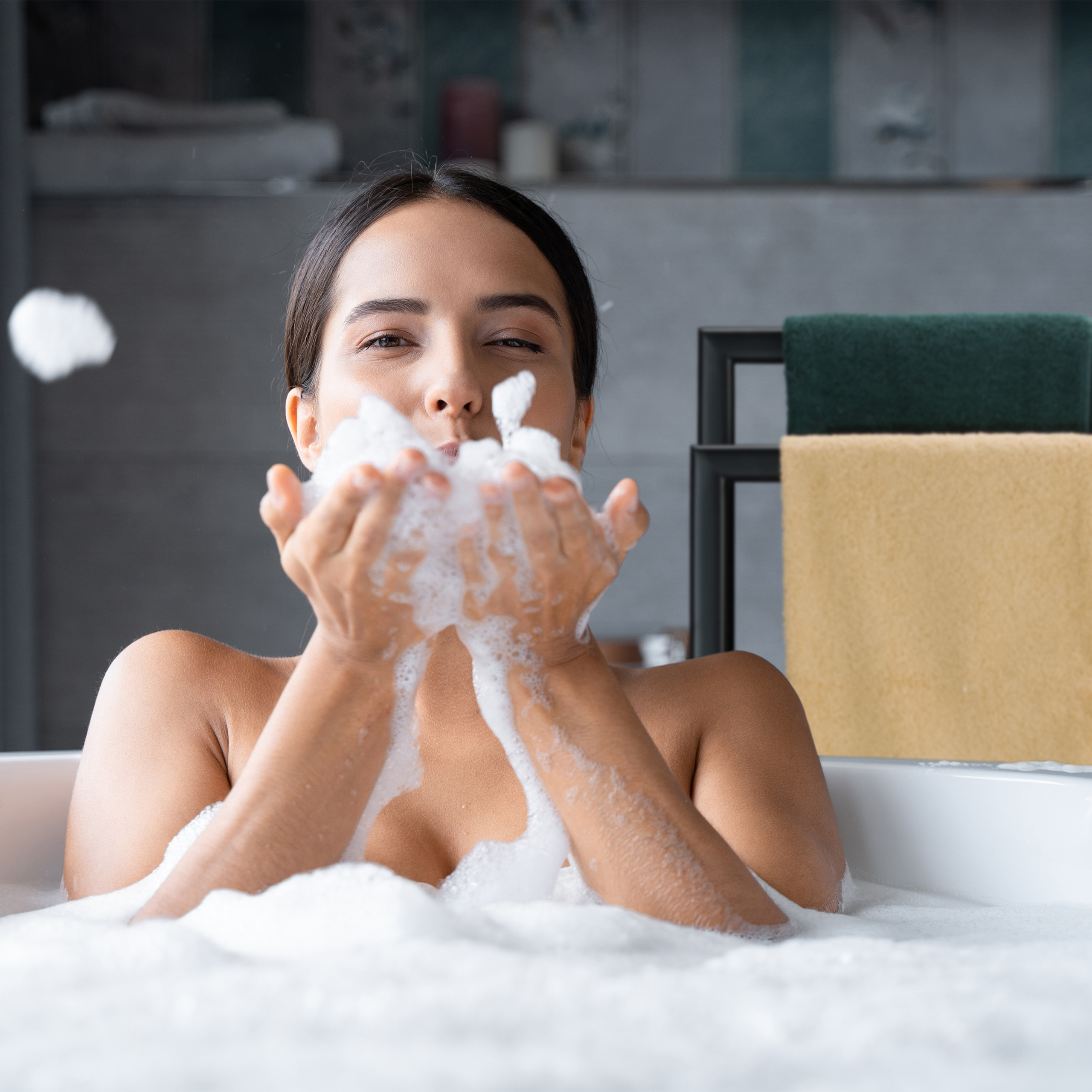 Eine Frau in der Badewanne bläst Seifenschaum von den Händen, in einem Badezimmer.