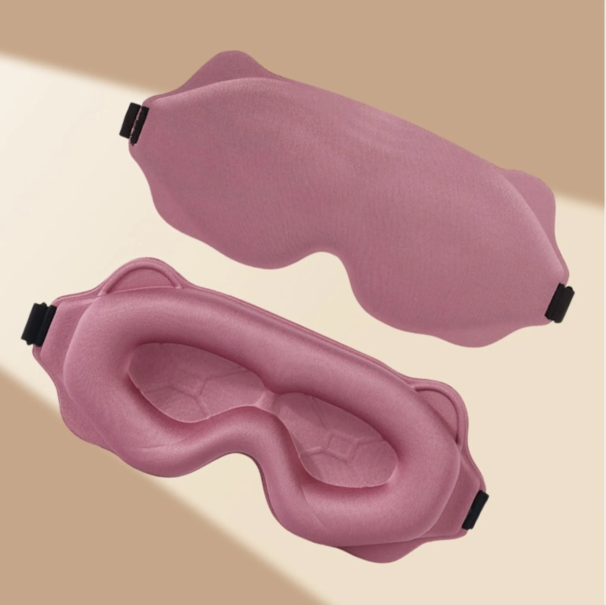 Zwei rosa geformte Schlafmasken mit schwarzen Riemen vor einem beigen geometrischen Hintergrund.