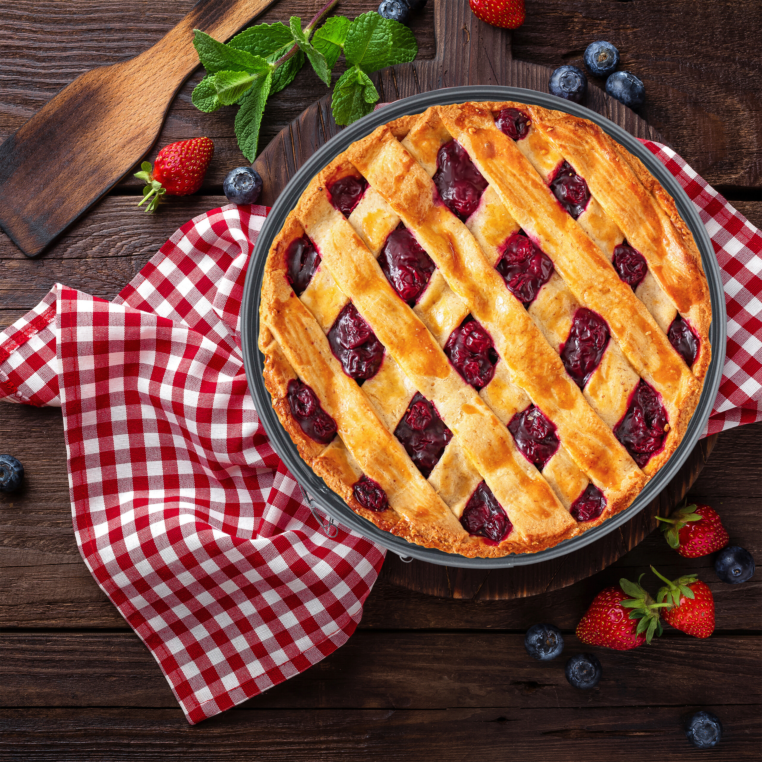 Ein Gitter-Kirschkuchen auf einem Holztisch mit rot-weißem Tuch, Erdbeeren, Blaubeeren und Minze.