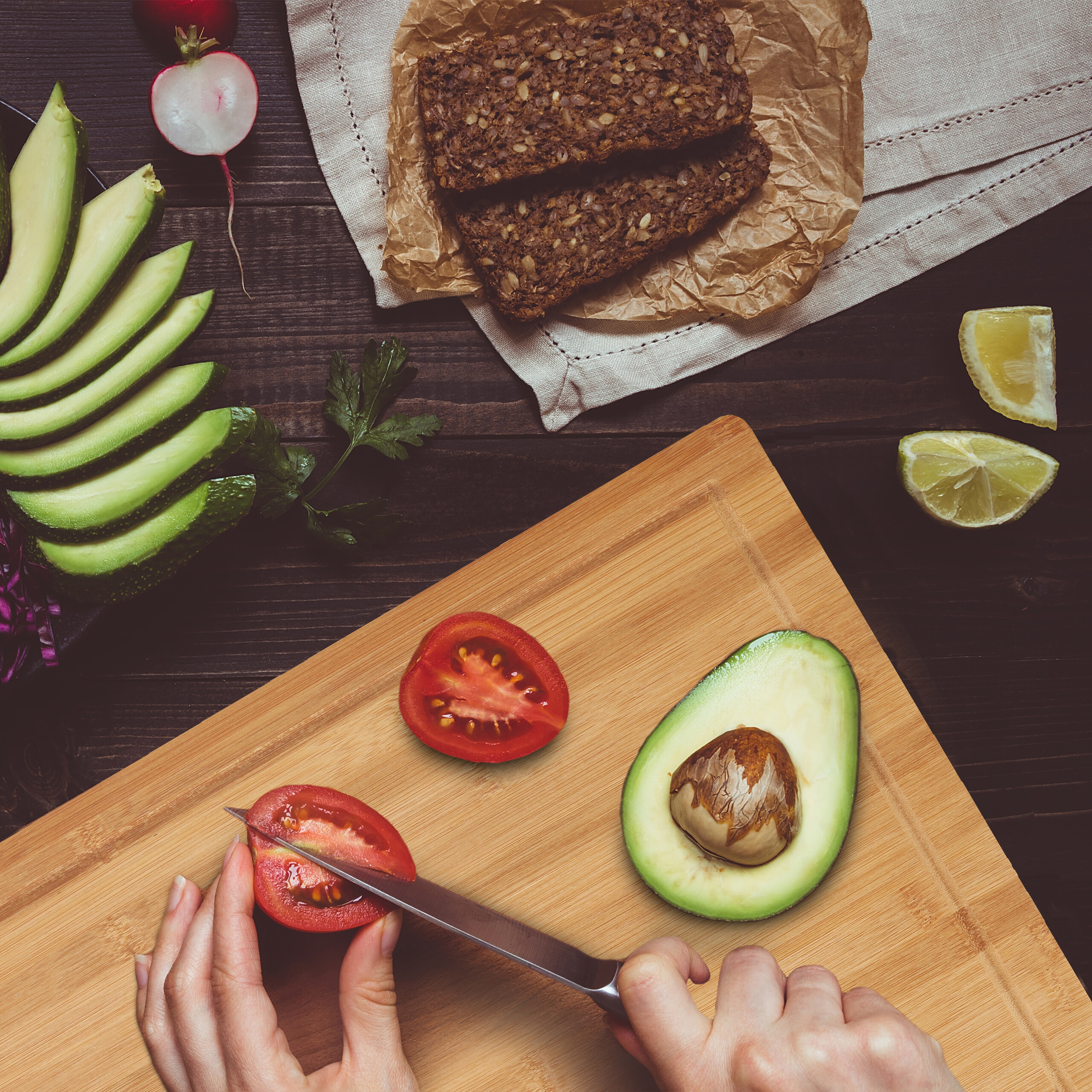 Mani tagliano un pomodoro su un tagliere con avocado, pane e verdure.