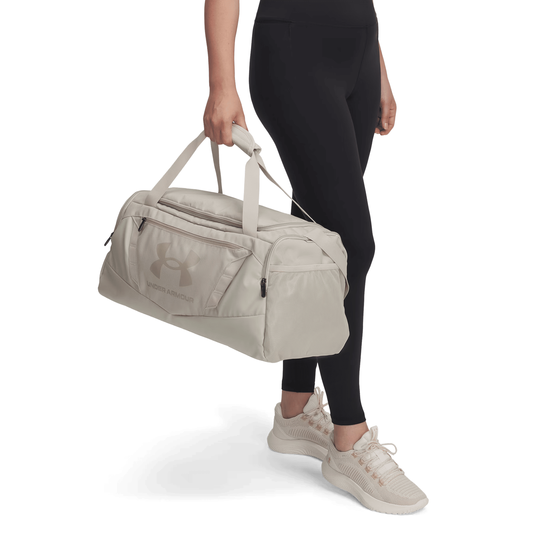 Person in schwarzen Leggings und hellen Turnschuhen hält eine helle Under Armour Sporttasche.