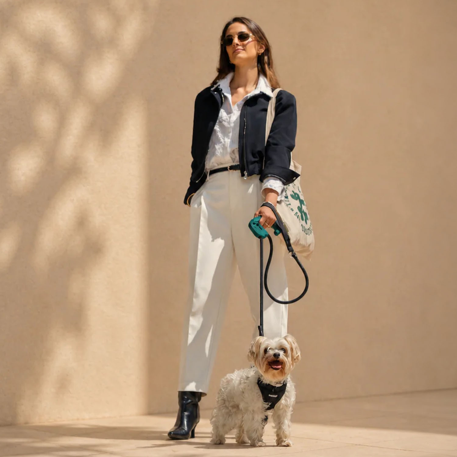 Una mujer con chaqueta negra y pantalones blancos está de pie contra una pared con un perro blanco con correa.