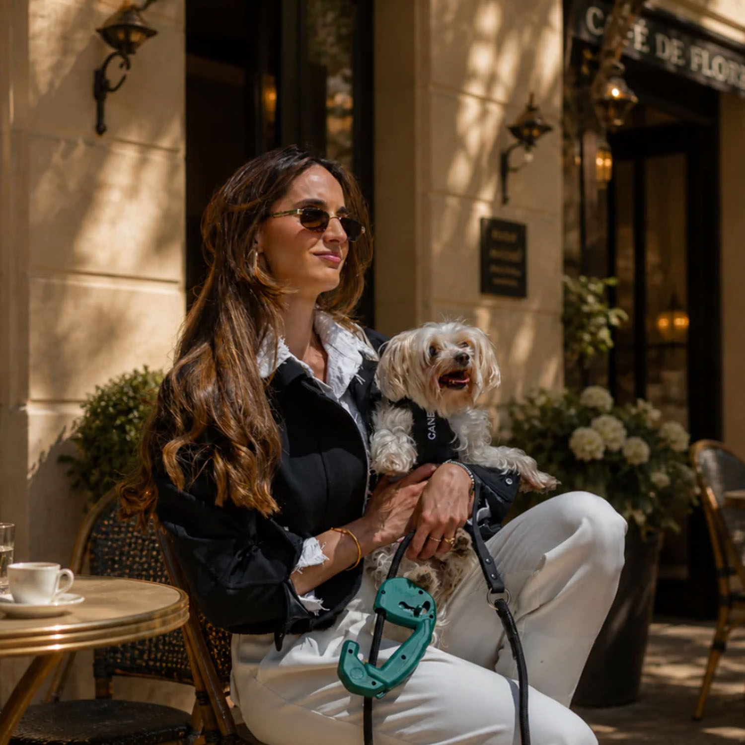 Una mujer de pelo castaño largo y gafas de sol sostiene a un perrito blanco en un café al aire libre.
