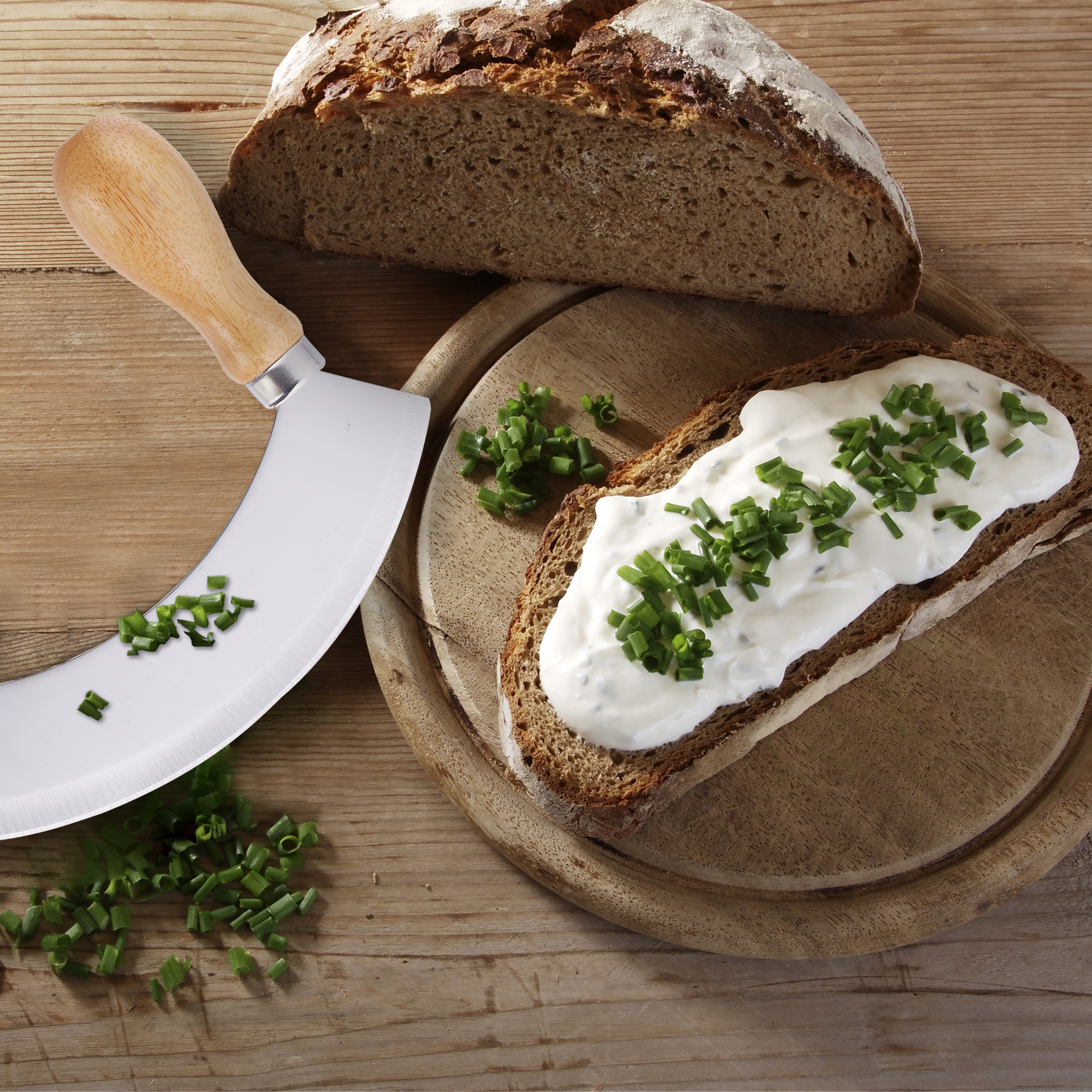 Dunkle Brotscheibe mit Frischkäse und Schnittlauch, daneben ein Messer und Brot auf einem Holzbrett.