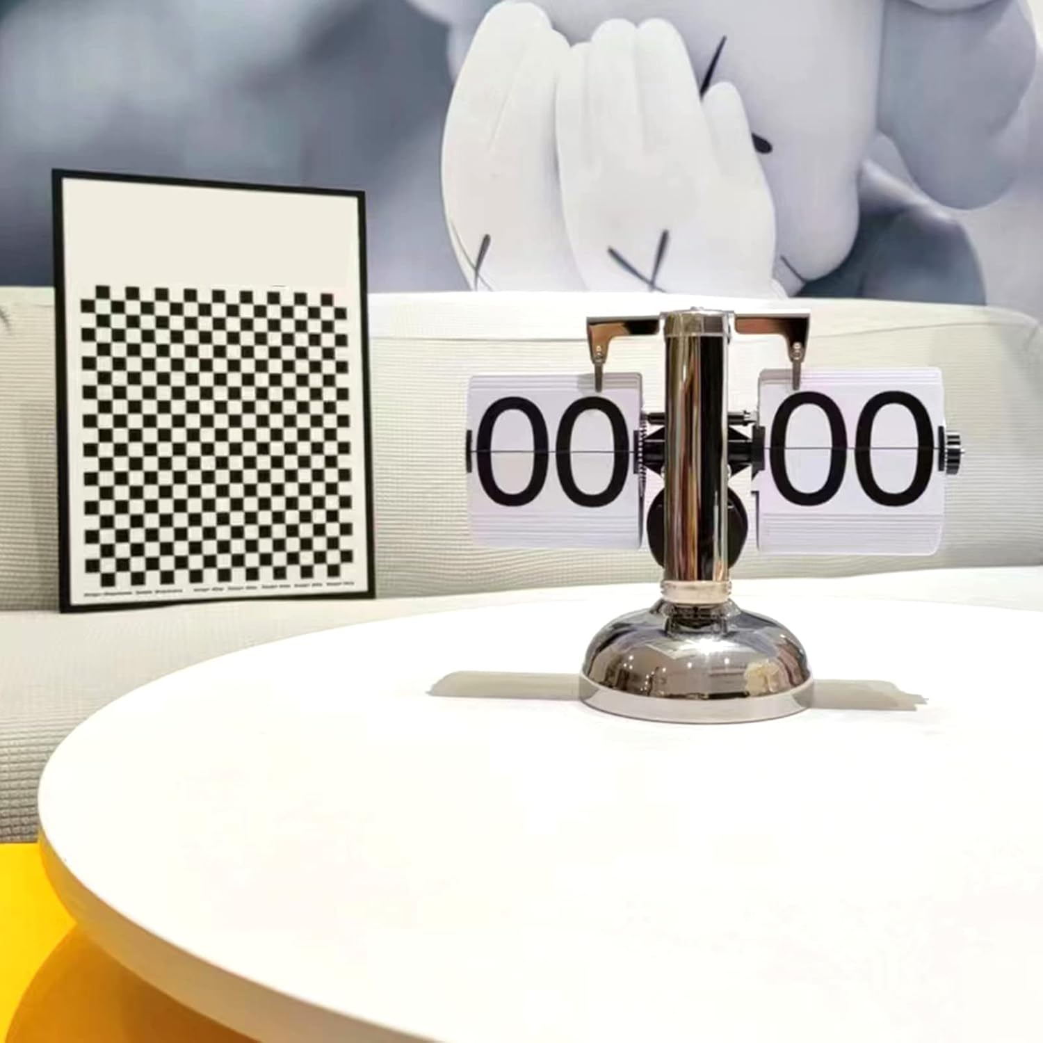 Eine silberne Flip-Uhr zeigt 00 00 auf einem weißen Tisch, mit karierter Kunst und einer weißen Figur im Hintergrund.