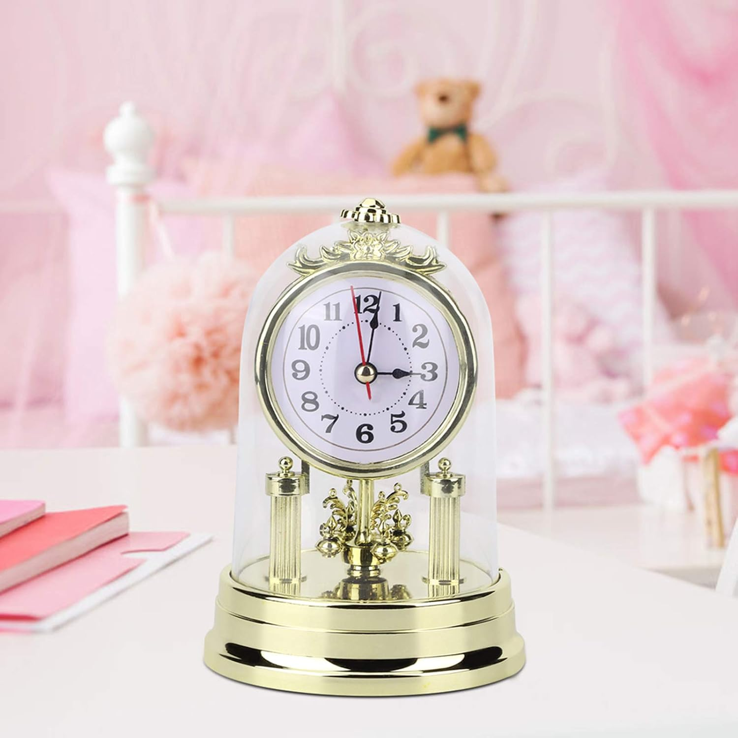 Eine goldene Standuhr unter einer Glasglocke auf einem weißen Tisch, im Hintergrund ein rosafarbenes Kinderzimmer.