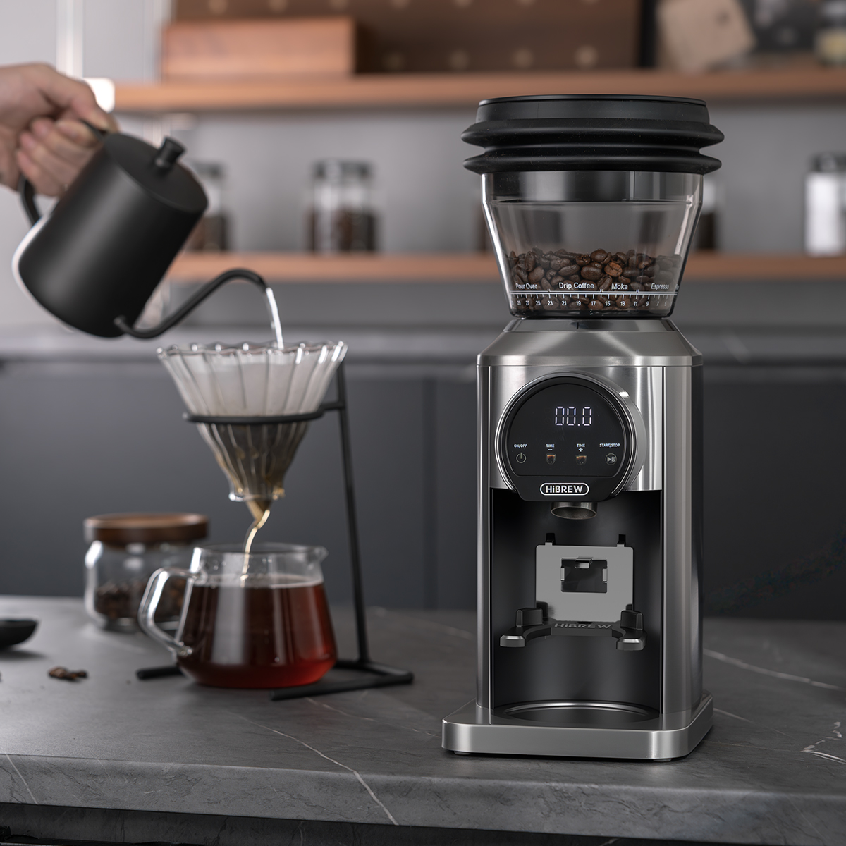 Eine elektrische Kaffeemühle steht auf einer Theke, während links Wasser aus einer Kanne in einen Pour-Over-Filter gegossen wird.