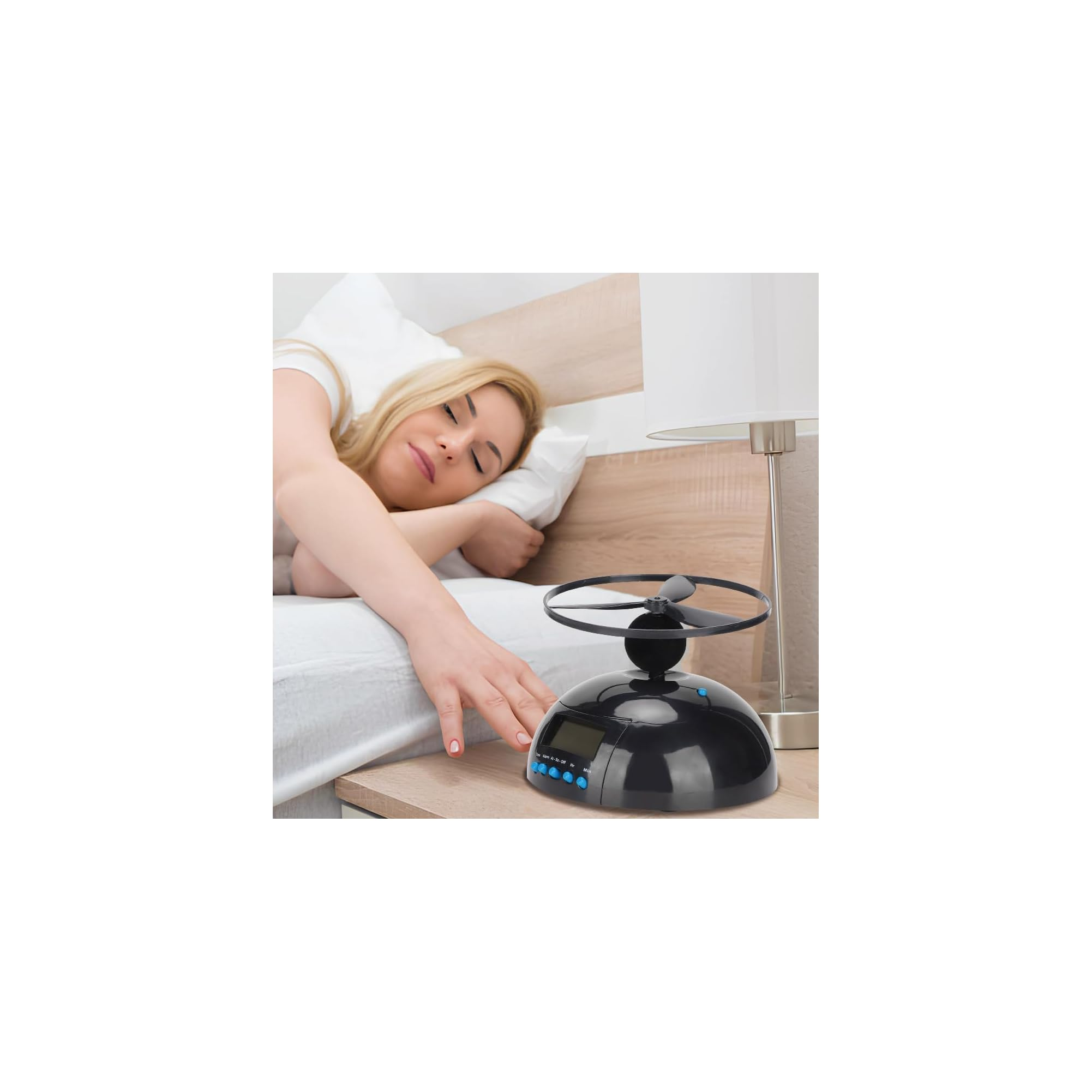 Eine blonde Frau im Bett schaltet eine schwarze, fliegende Weckuhr auf dem Nachttisch aus.