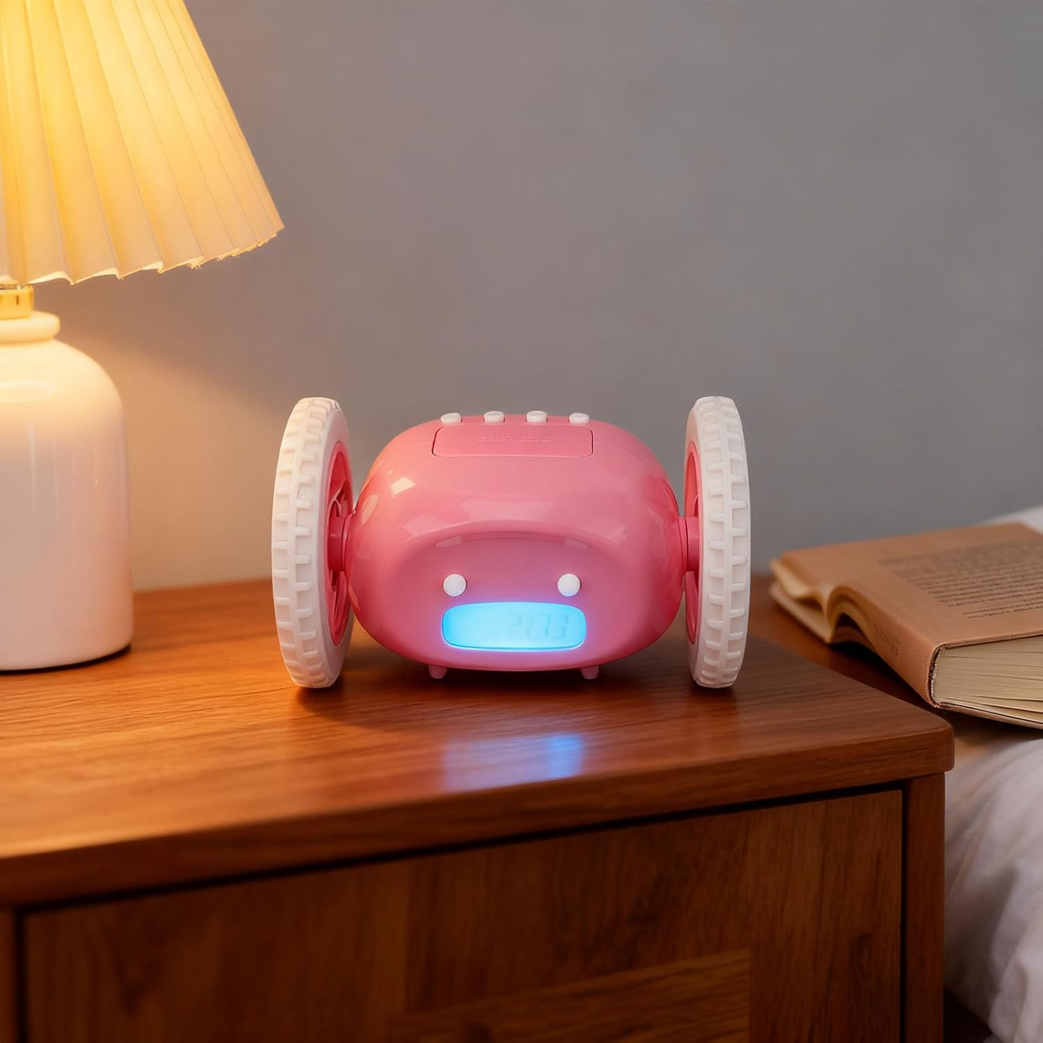 Ein rosafarbener roboterartiger Digitalwecker mit großen weißen Rädern steht auf einem Nachttisch neben Lampe und Buch.