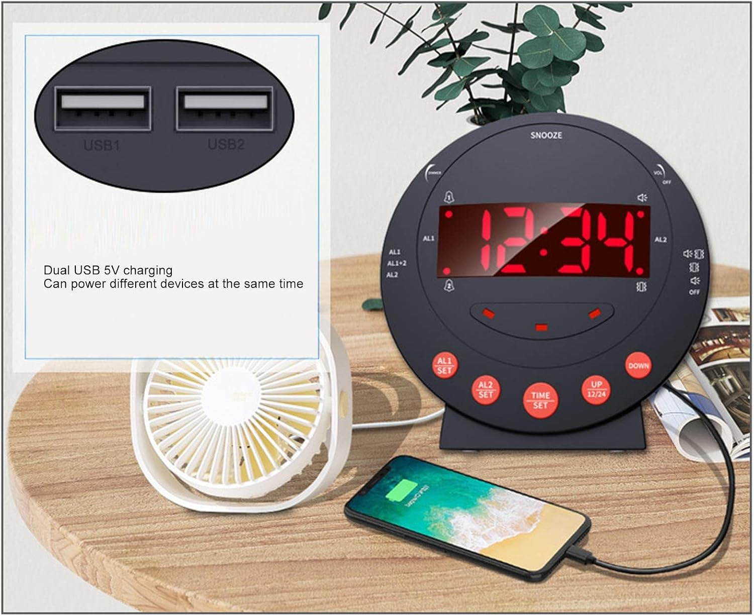 Ein runder digitaler Wecker mit zwei USB-Anschlüssen lädt ein Smartphone und einen Ventilator auf einem Holztisch.