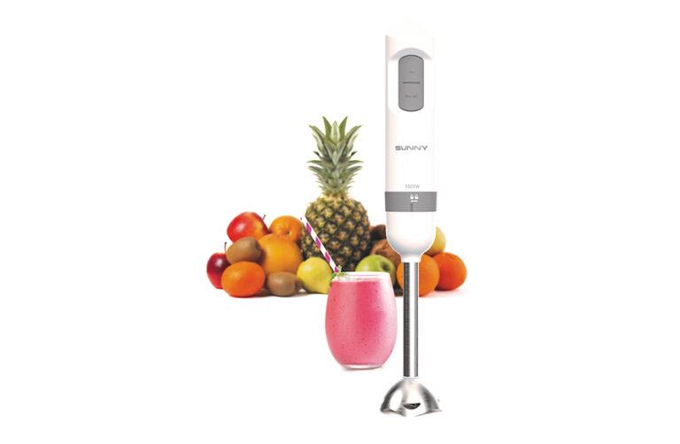 Beyaz bir el blenderi, pembe bir smoothie bardağı ve ananas, elma, portakal gibi çeşitli meyvelerin önünde duruyor.