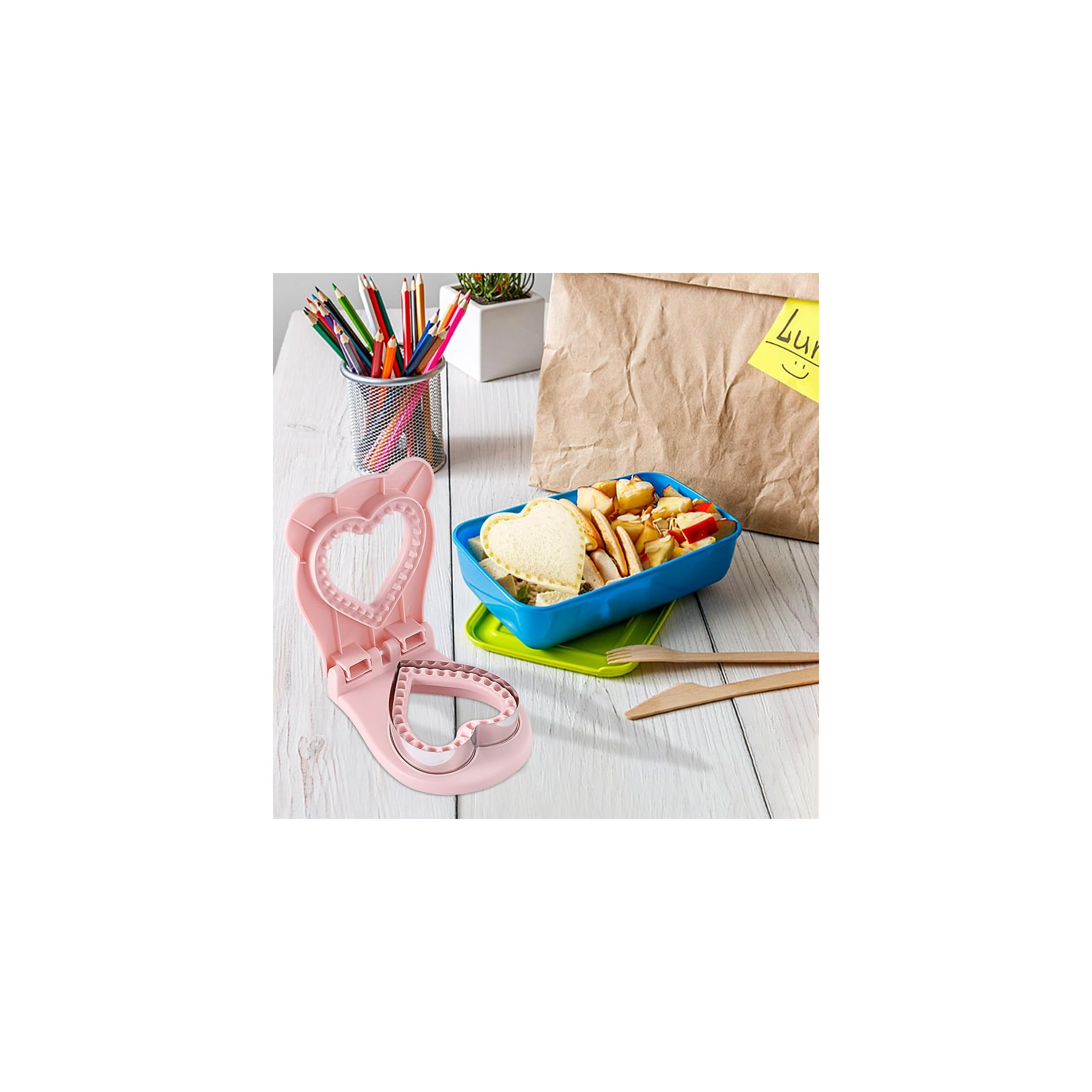 Rosa herzförmige Sandwichpresse auf Holztisch neben blauer Brotdose und Papiertüte.