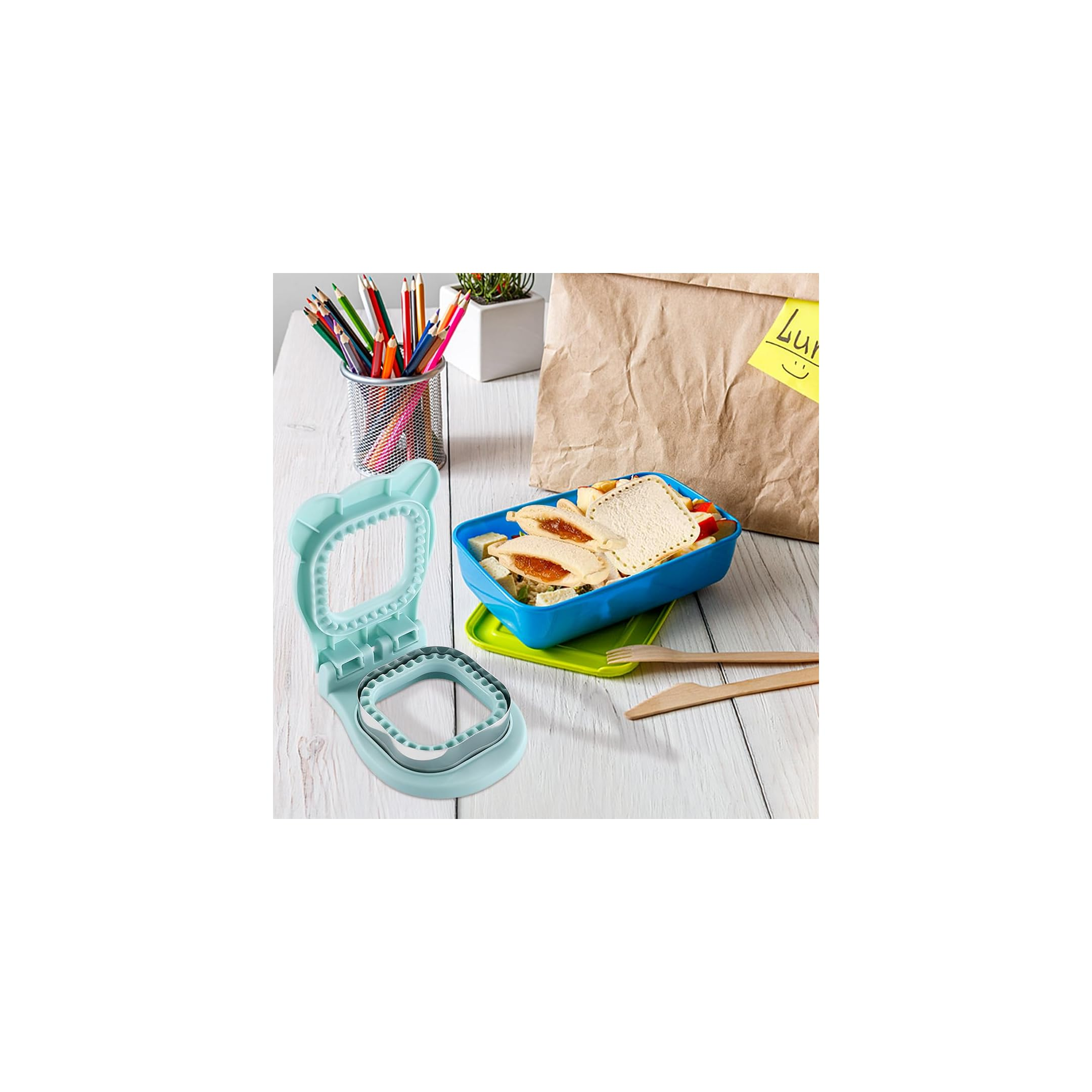 Blaue Lunchbox mit Sandwich, türkisfarbener Sandwich-Cutter und Papiertüte auf weißem Holz.