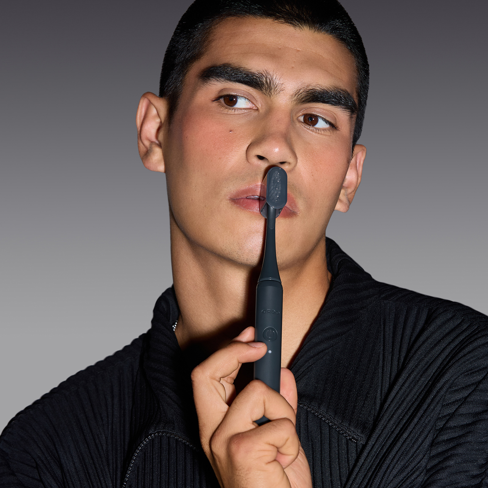Un hombre con camisa negra sostiene un cepillo eléctrico oscuro verticalmente cerca de sus labios contra fondo gris.